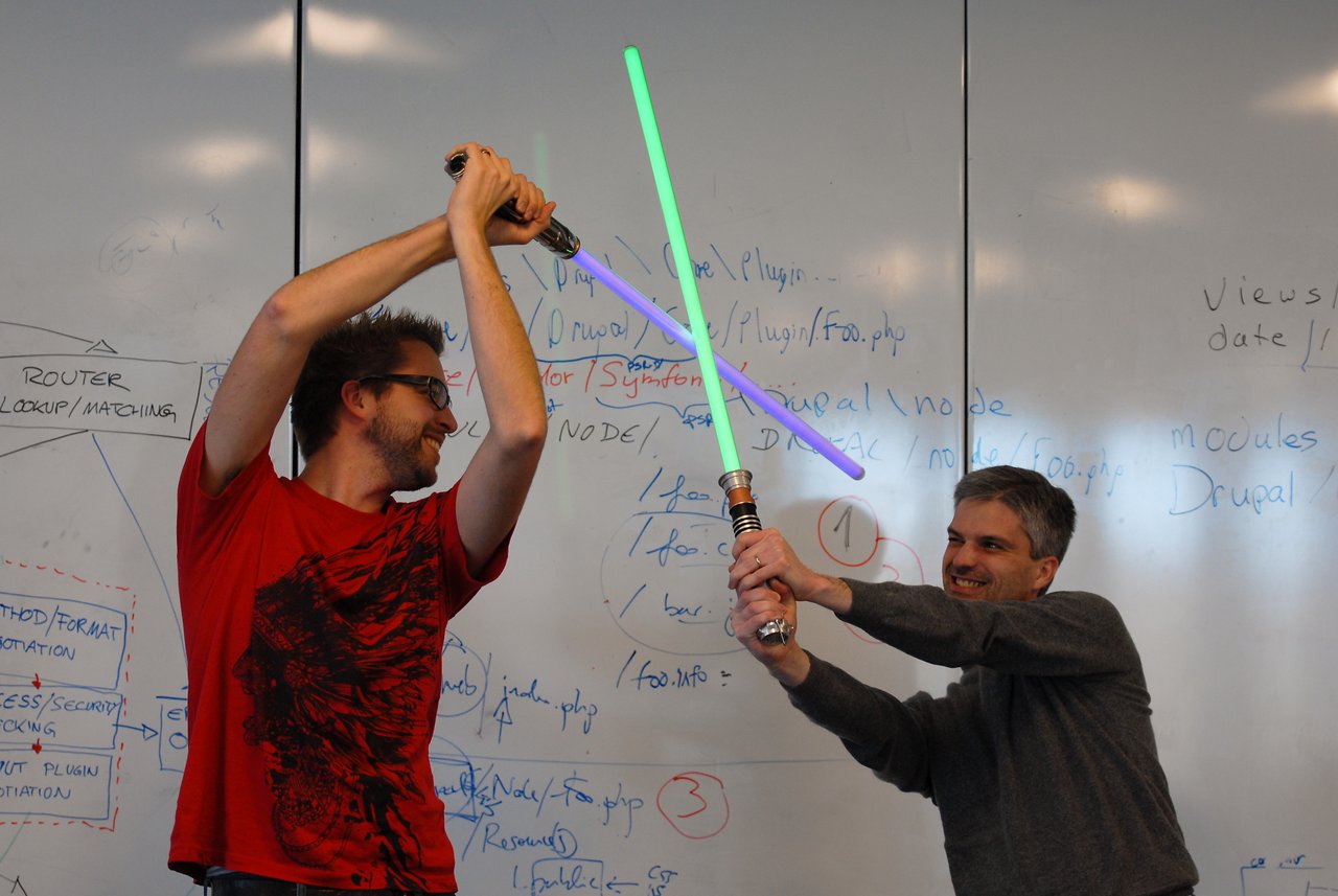 Two people smiling and playfully dueling with toy lightsabers in front of a whiteboard filled with notes.