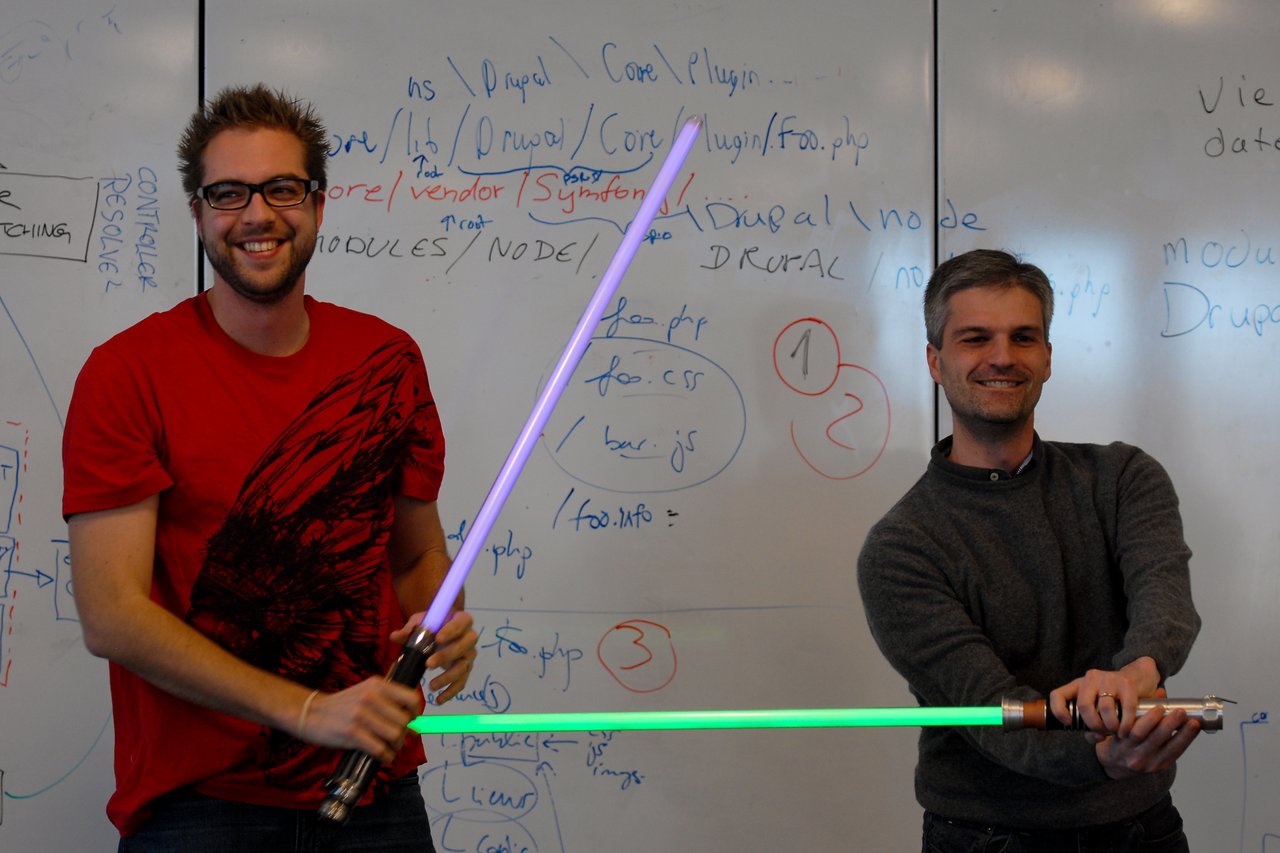 Two smiling people hold toy lightsabers in front of a whiteboard covered in handwritten notes and diagrams.