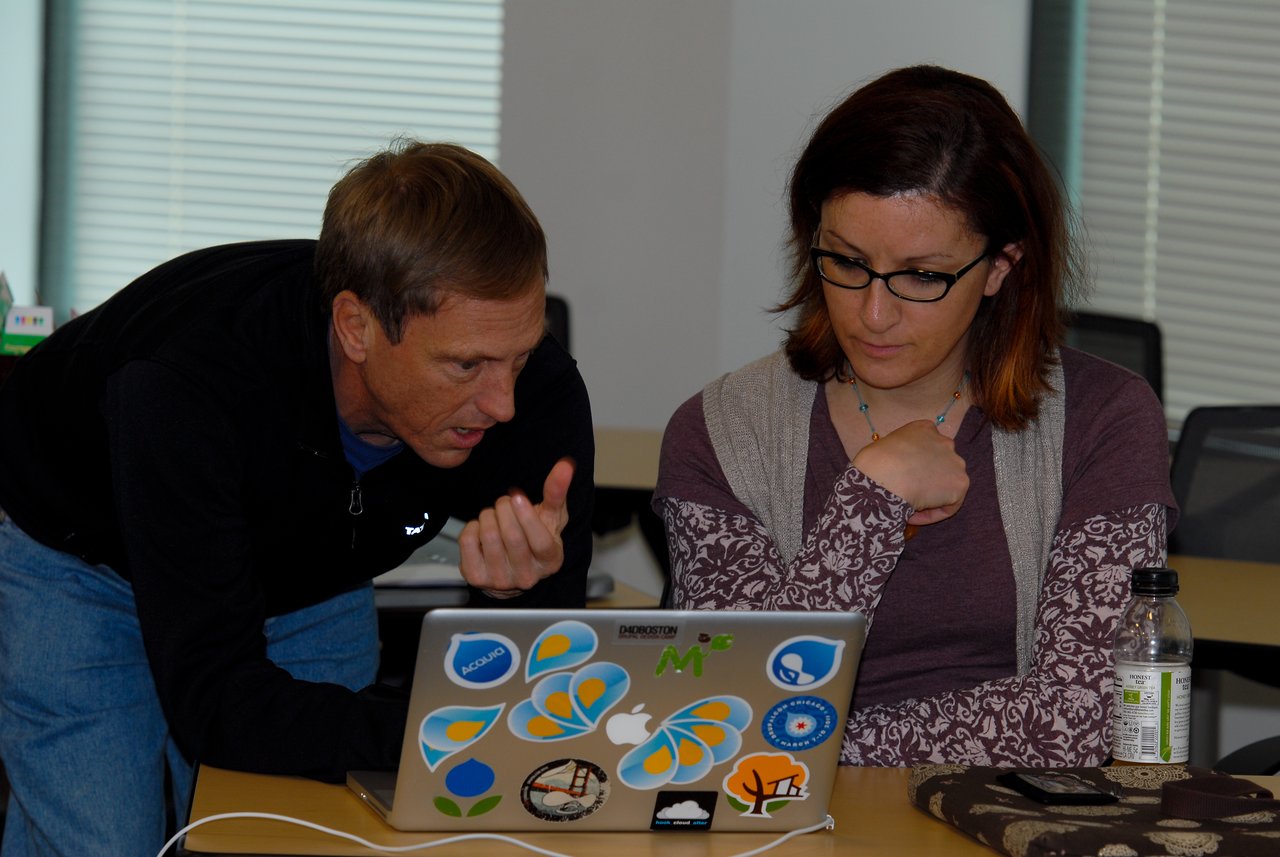 A man leans over and explains something to a woman focused on her laptop during a work session.