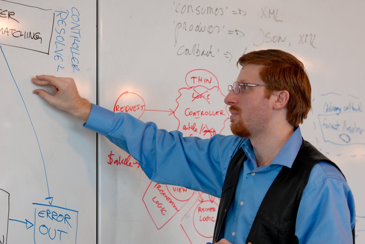 A man in a blue shirt points at a whiteboard filled with diagrams and notes during a discussion.