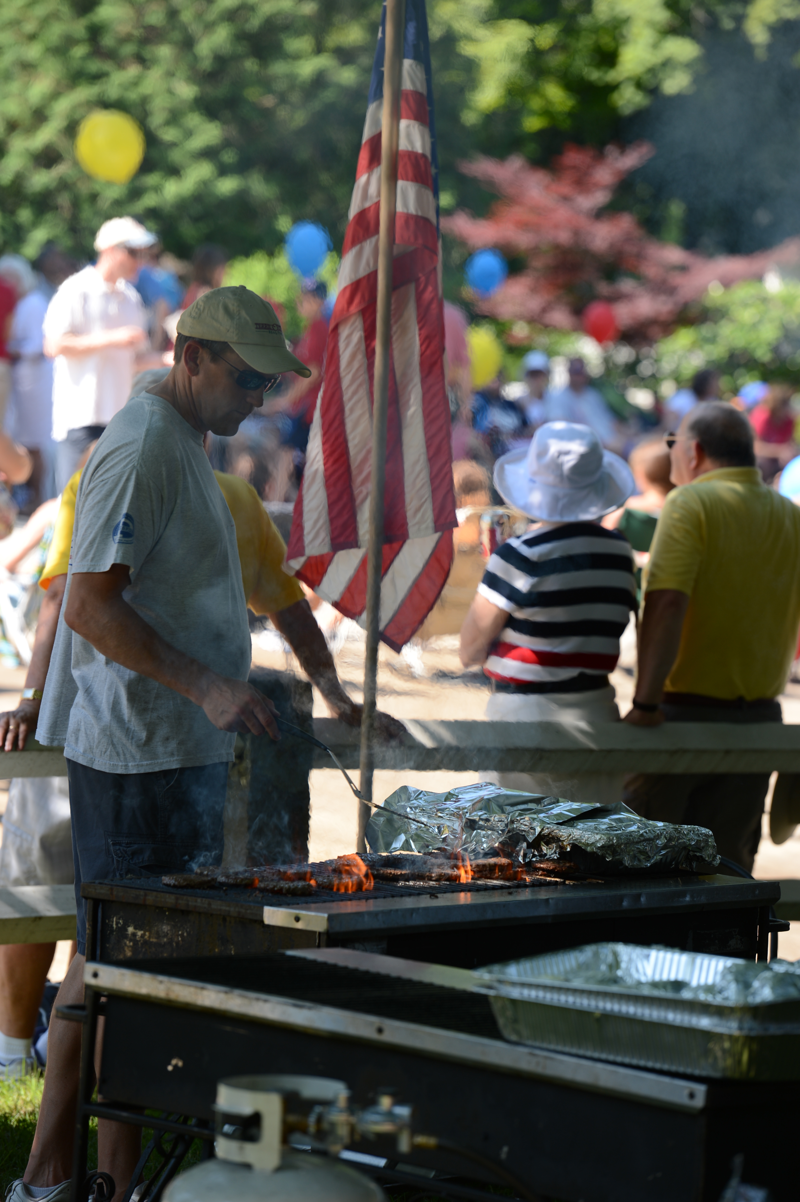 A man grills food on an open flame at an outdoor gathering, with an American flag nearby.