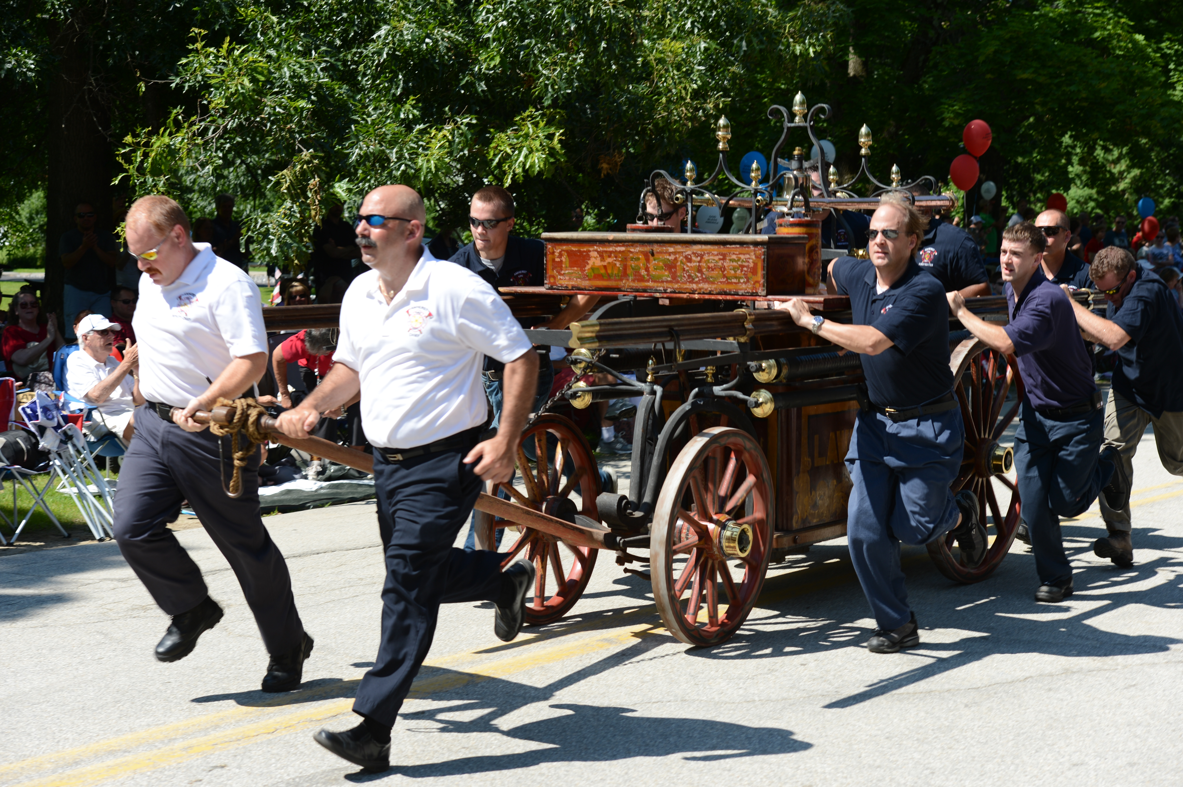 A group of people in uniform push and pull an antique fire wagon during a parade.