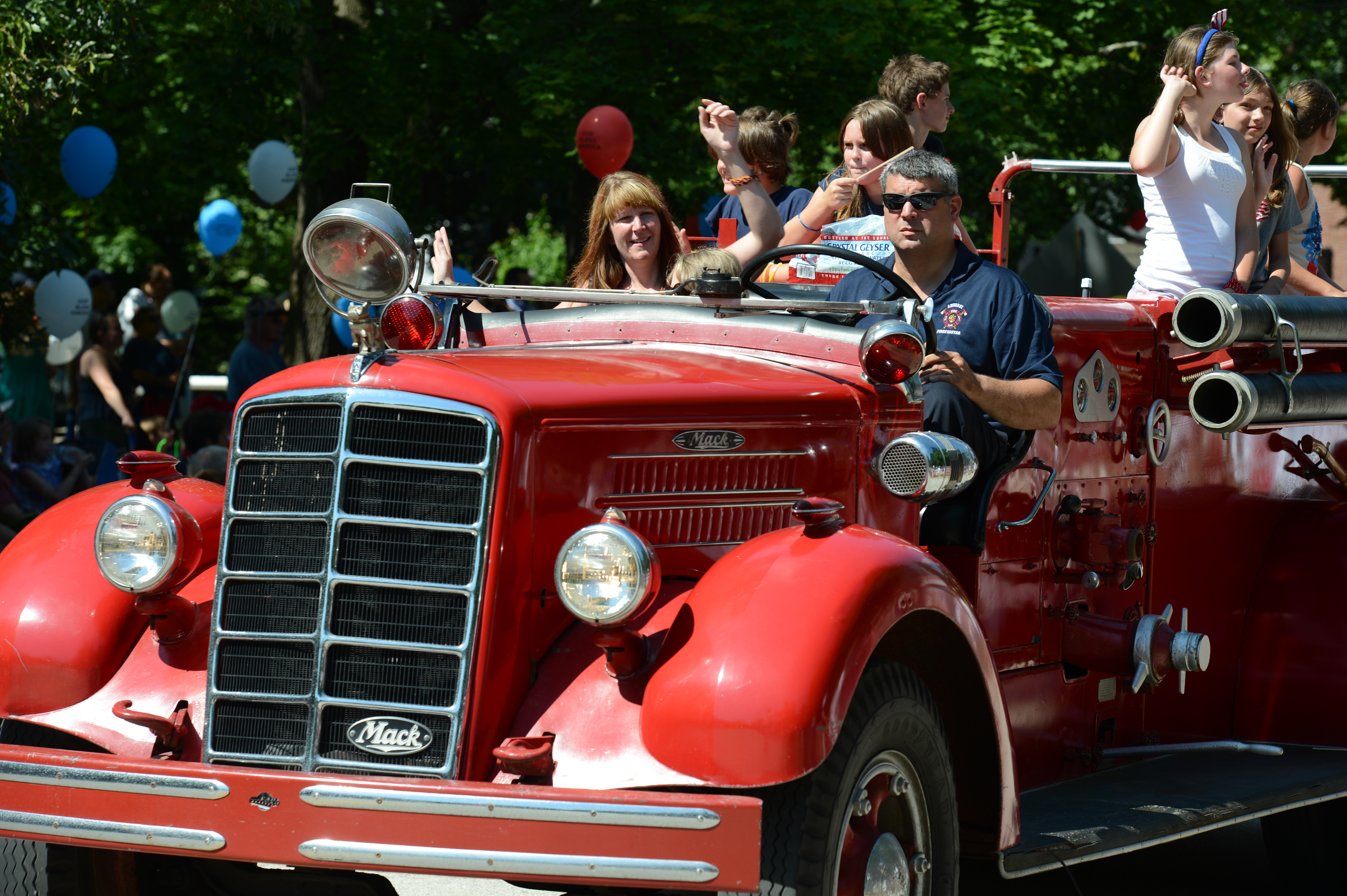 A red vintage fire truck drives in a parade, with people on board waving to the crowd.