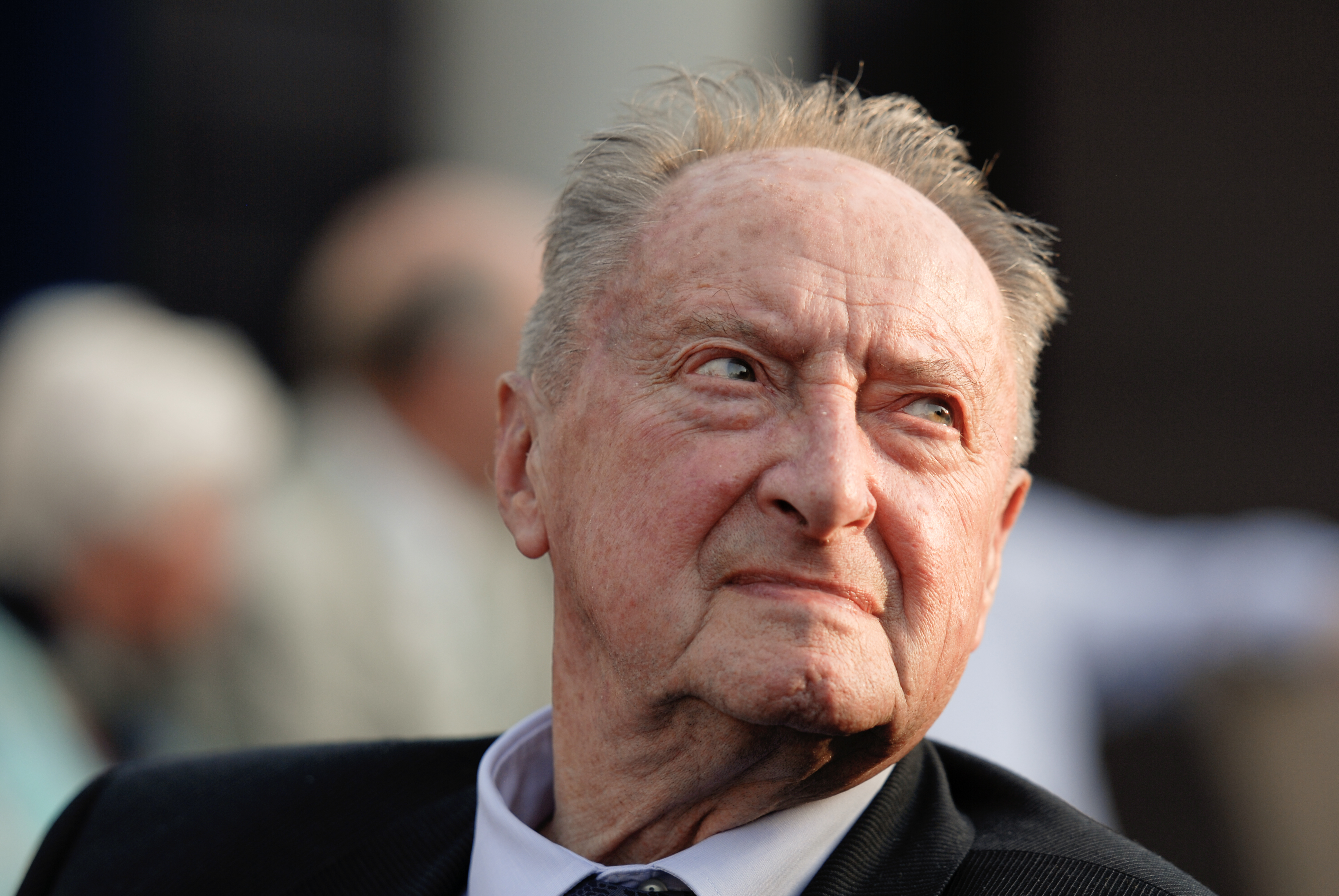 An elderly man in a suit looks off to the side at a gathering, with blurred people in the background.