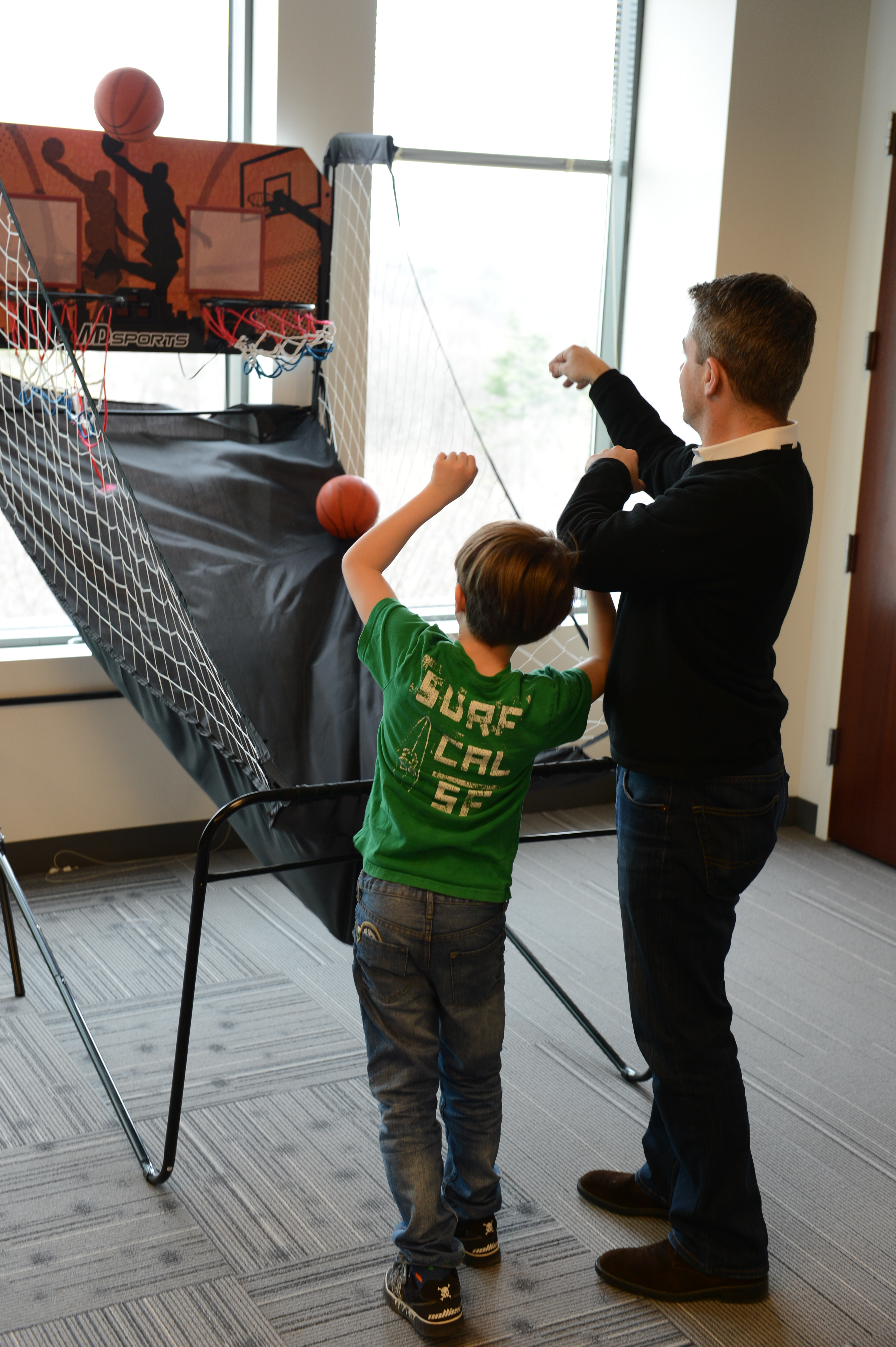 An adult and child play an indoor basketball arcade game, both shooting the ball toward the hoop.