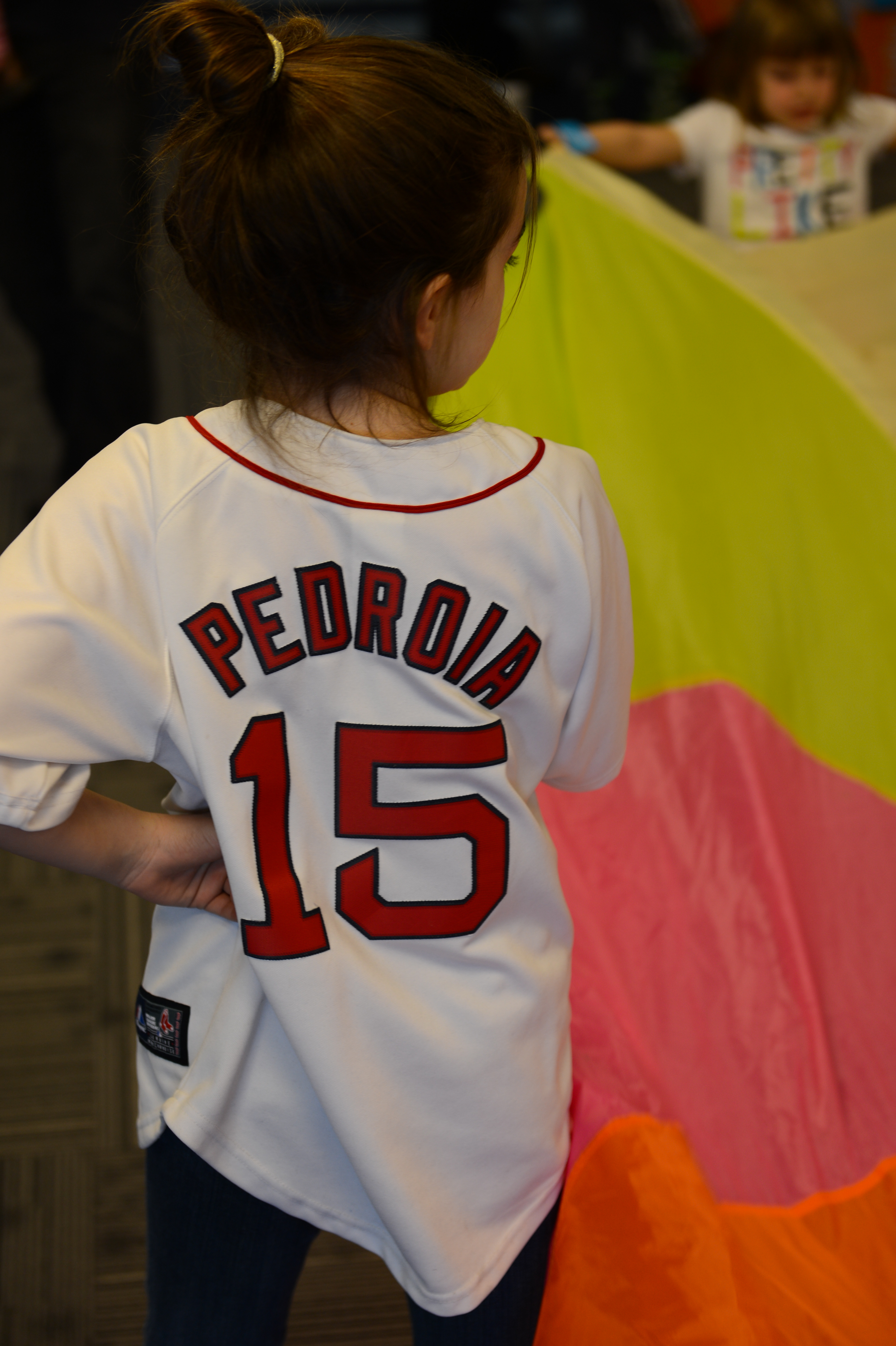 A child wearing a baseball jersey stands near a colorful parachute while another child plays in the background.