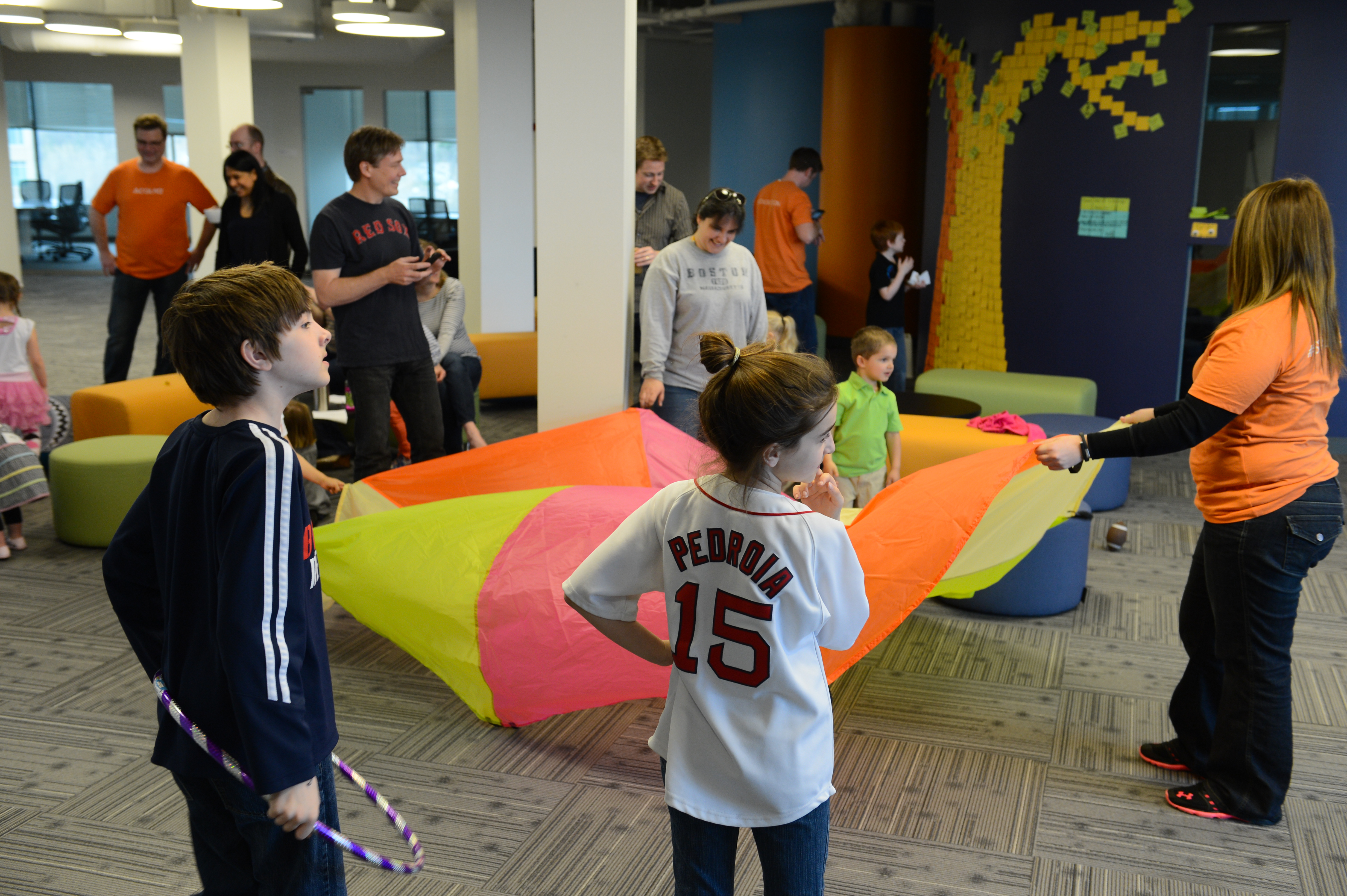 Children and adults play with a colorful parachute in an office space during a "Bring Your Kids to Work" event.