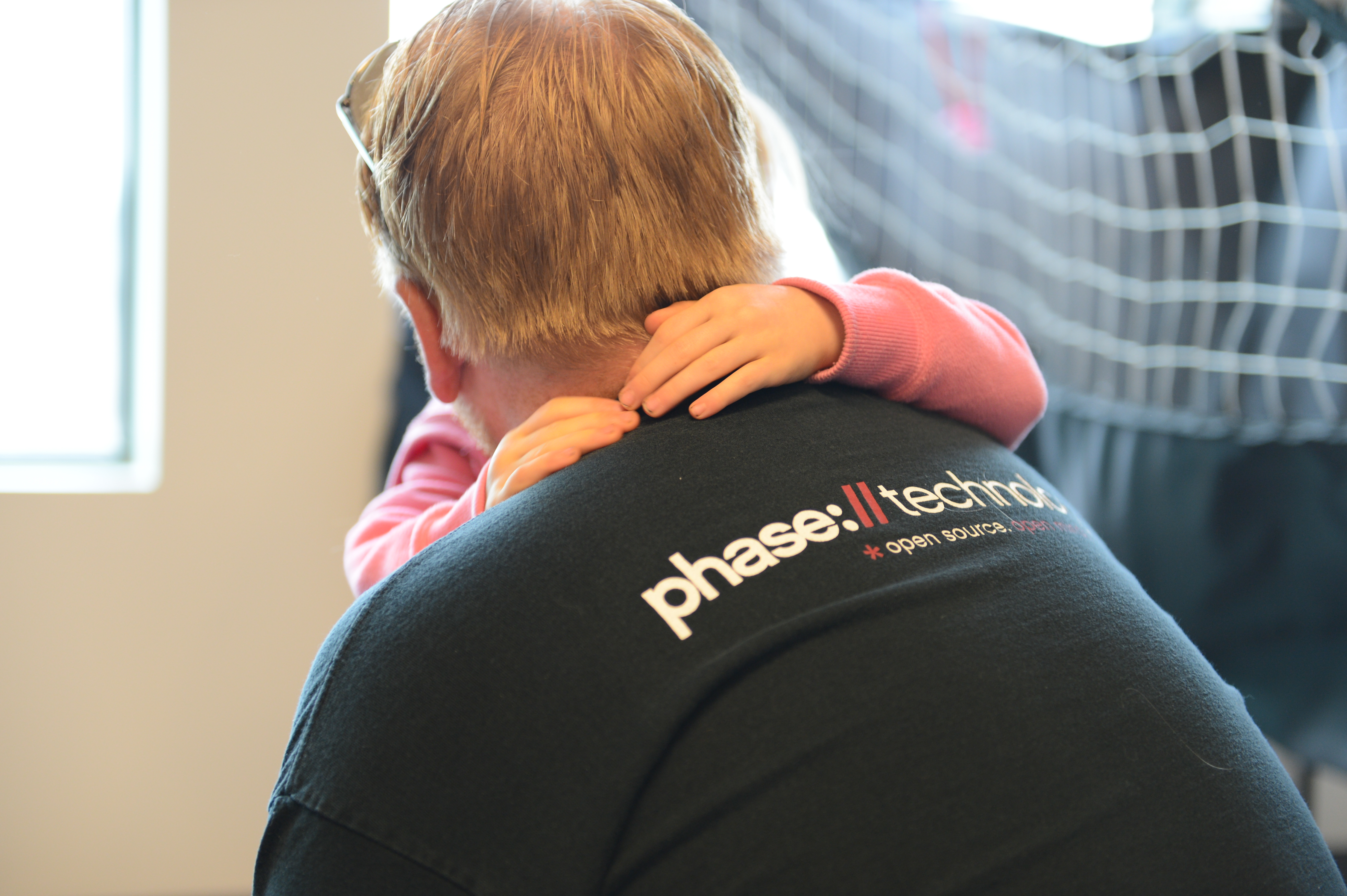 A child in a pink sweater hugs an adult wearing a black "Phase II Technology" shirt from behind.
