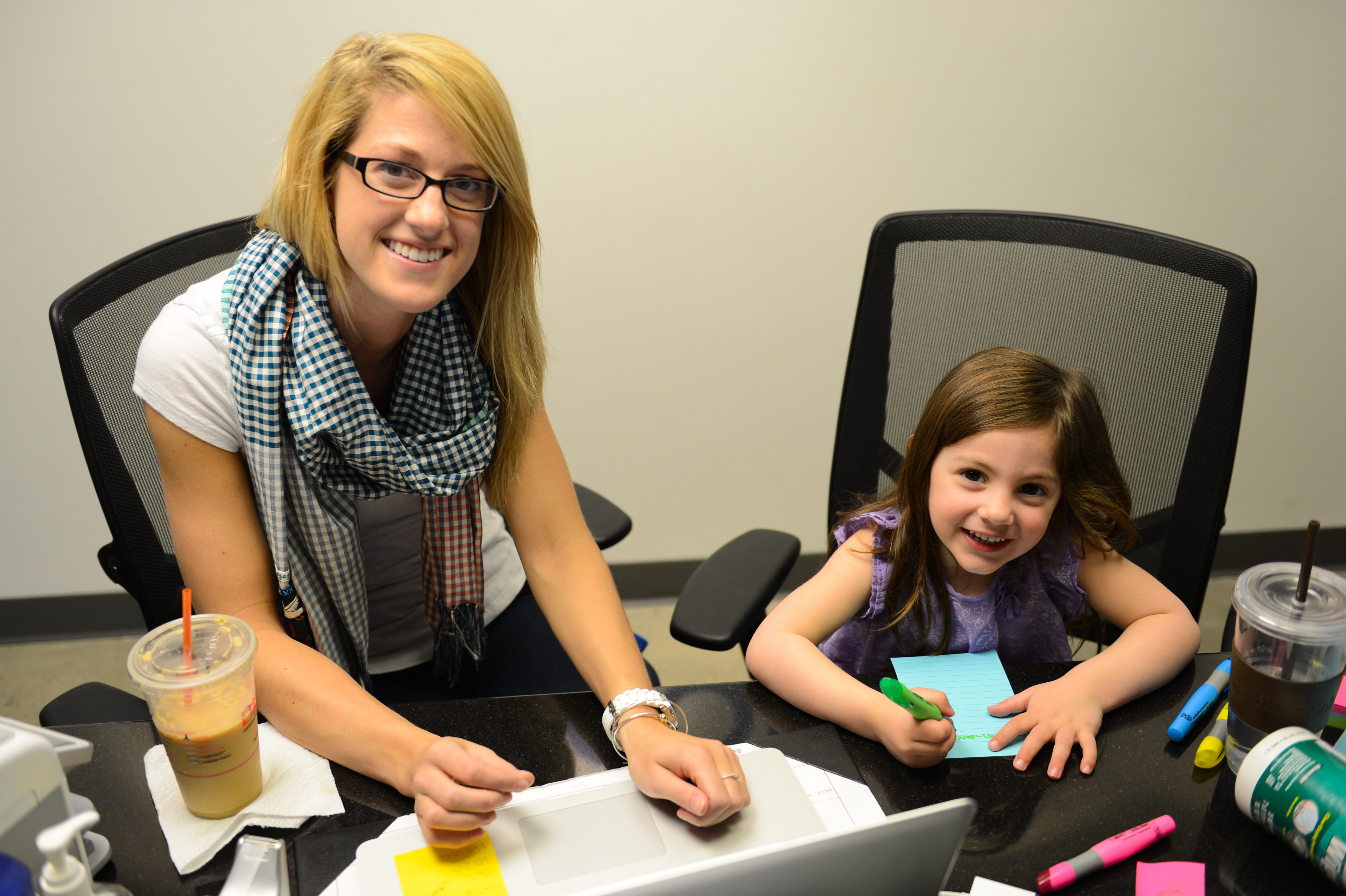 A woman and a young girl sit at a desk, smiling while the girl writes on a notepad with markers.
