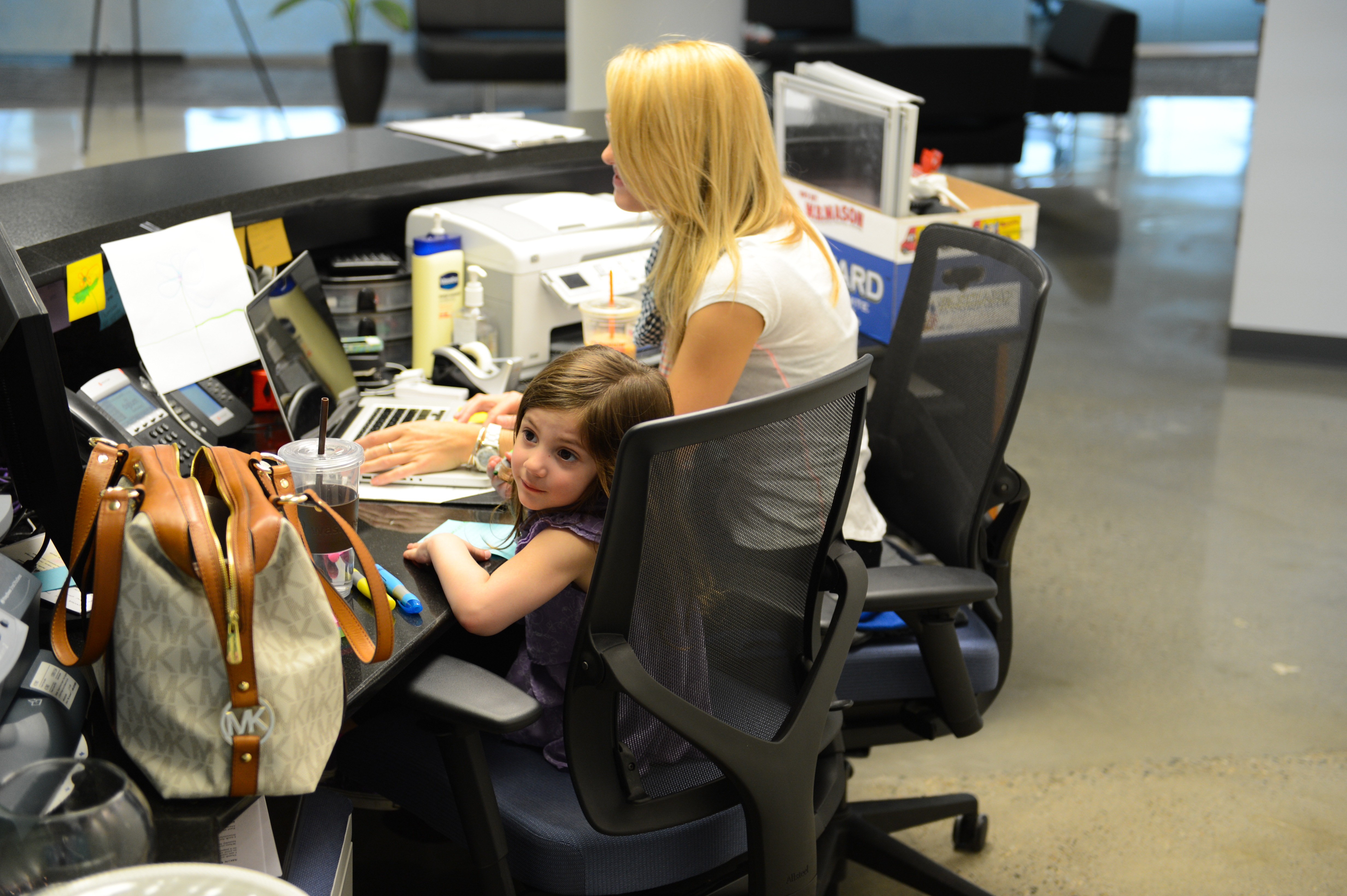 A woman works on a laptop at a desk while a young girl sits beside her, looking at the camera.