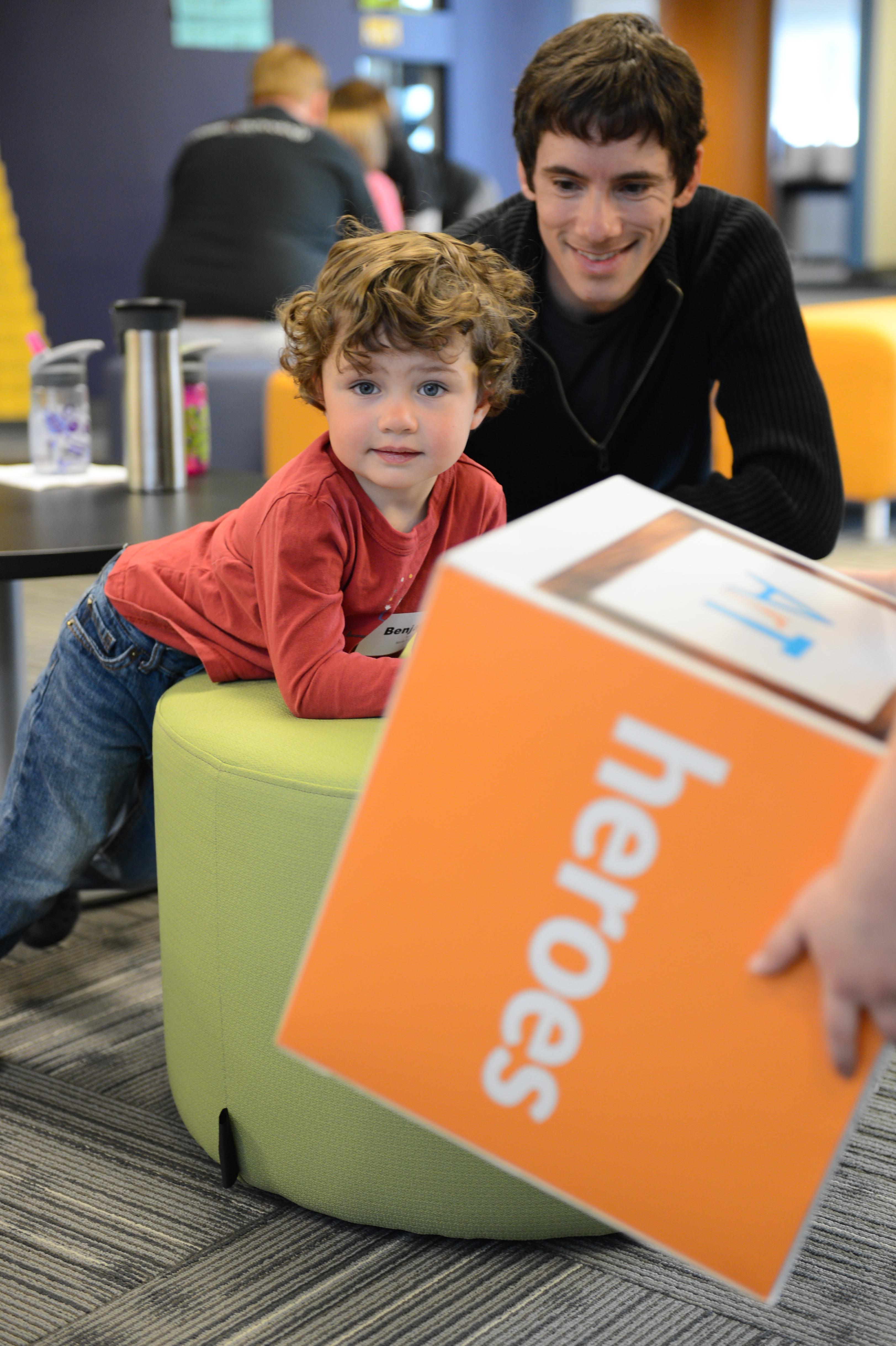 A child leans on a green stool while reaching for an orange box labeled "heroes", with an adult smiling nearby.