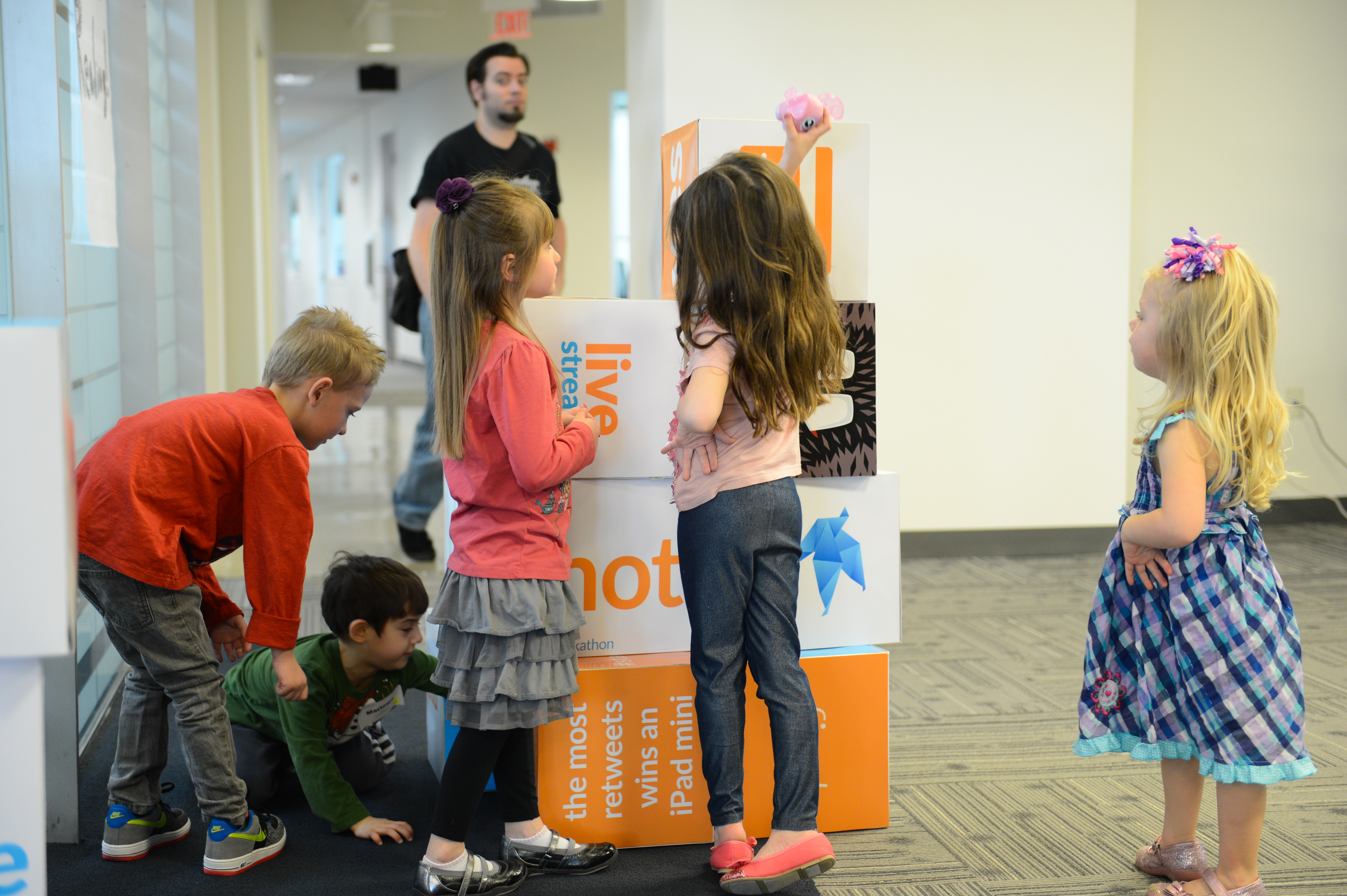 Several children gather around stacked boxes, with one reaching to place a small toy on top.
