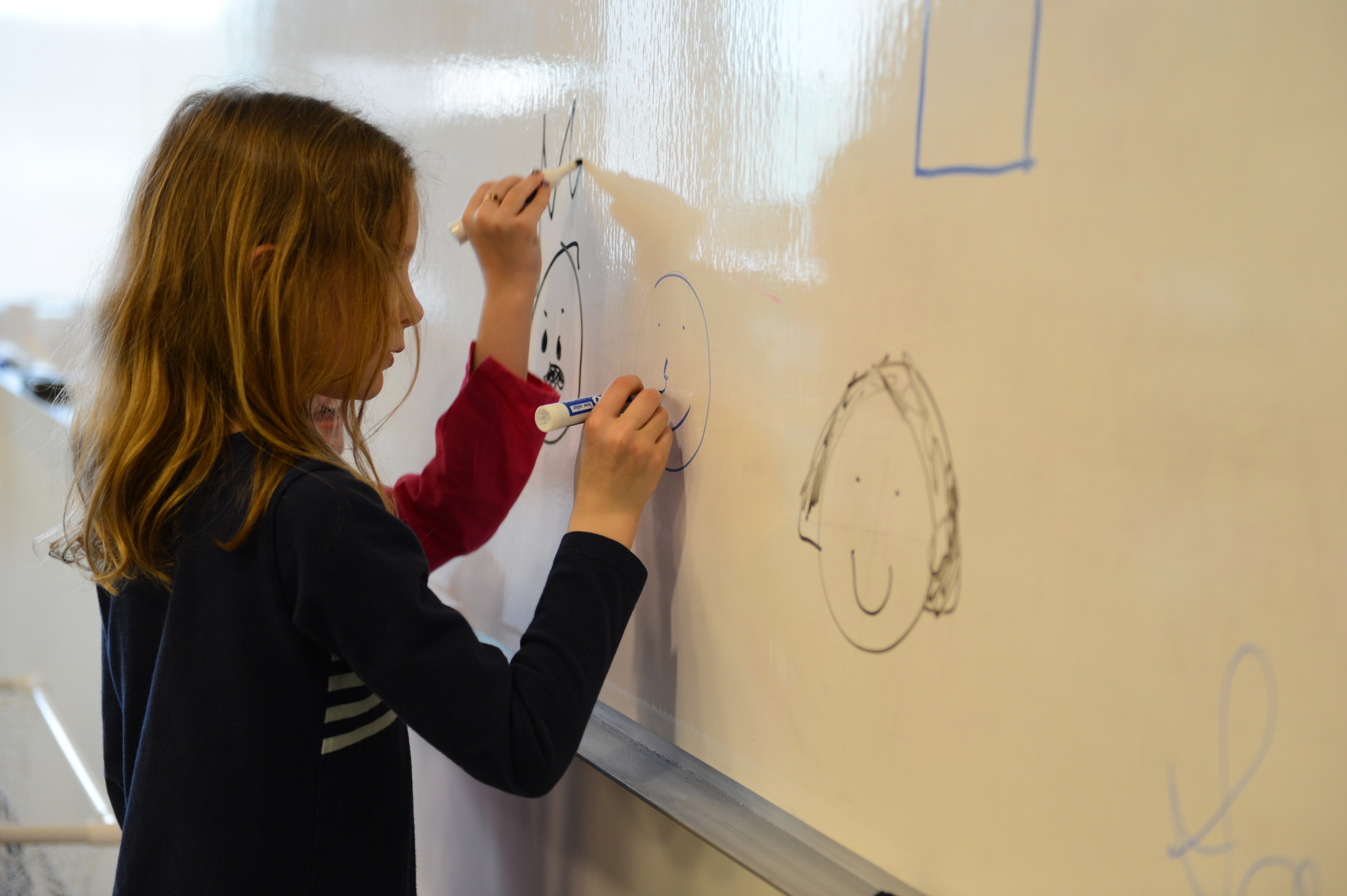 Two children draw faces on a whiteboard using markers.