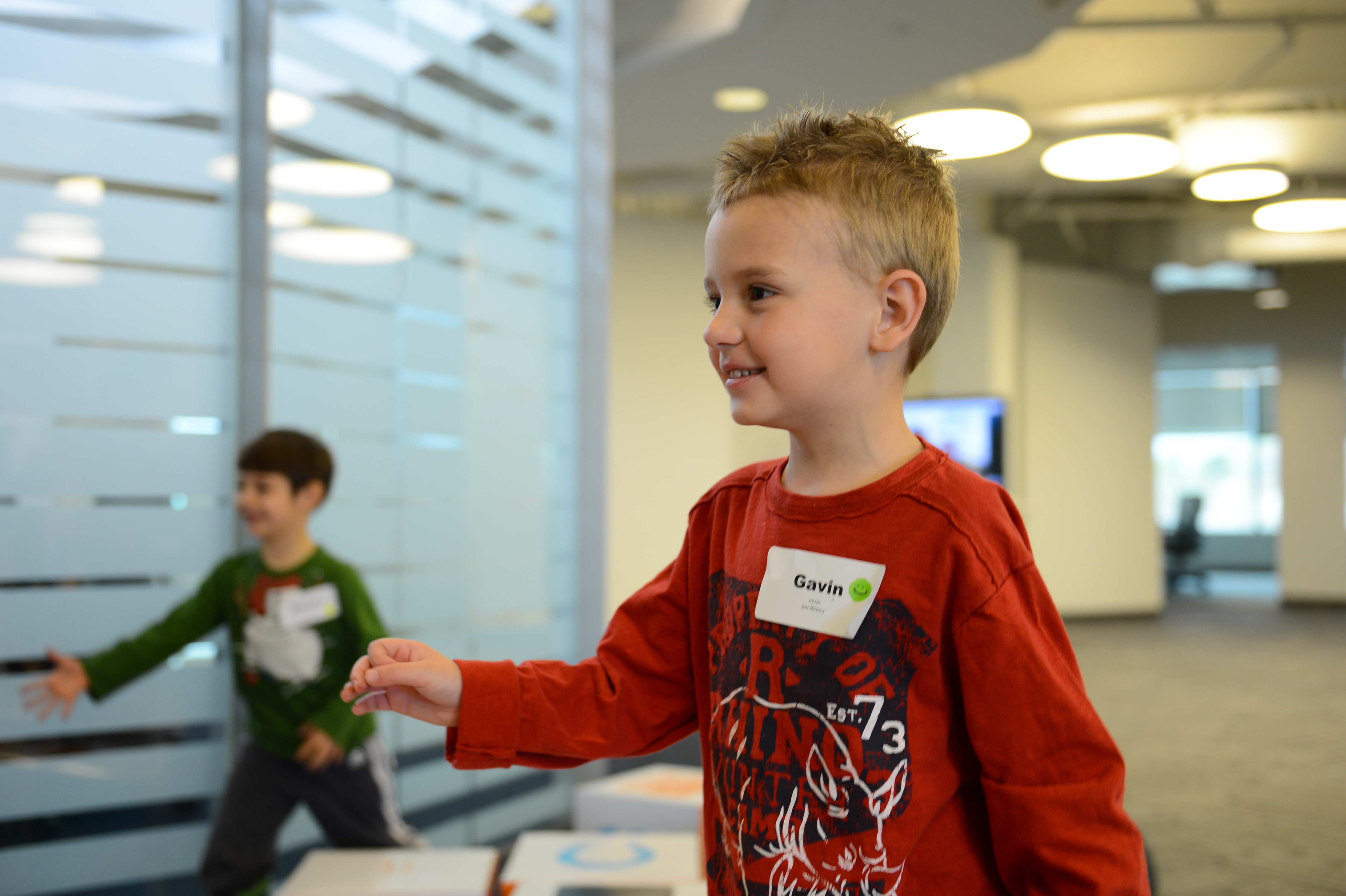 A young boy wearing a name tag smiles while playing indoors, with another child reaching out in the background.