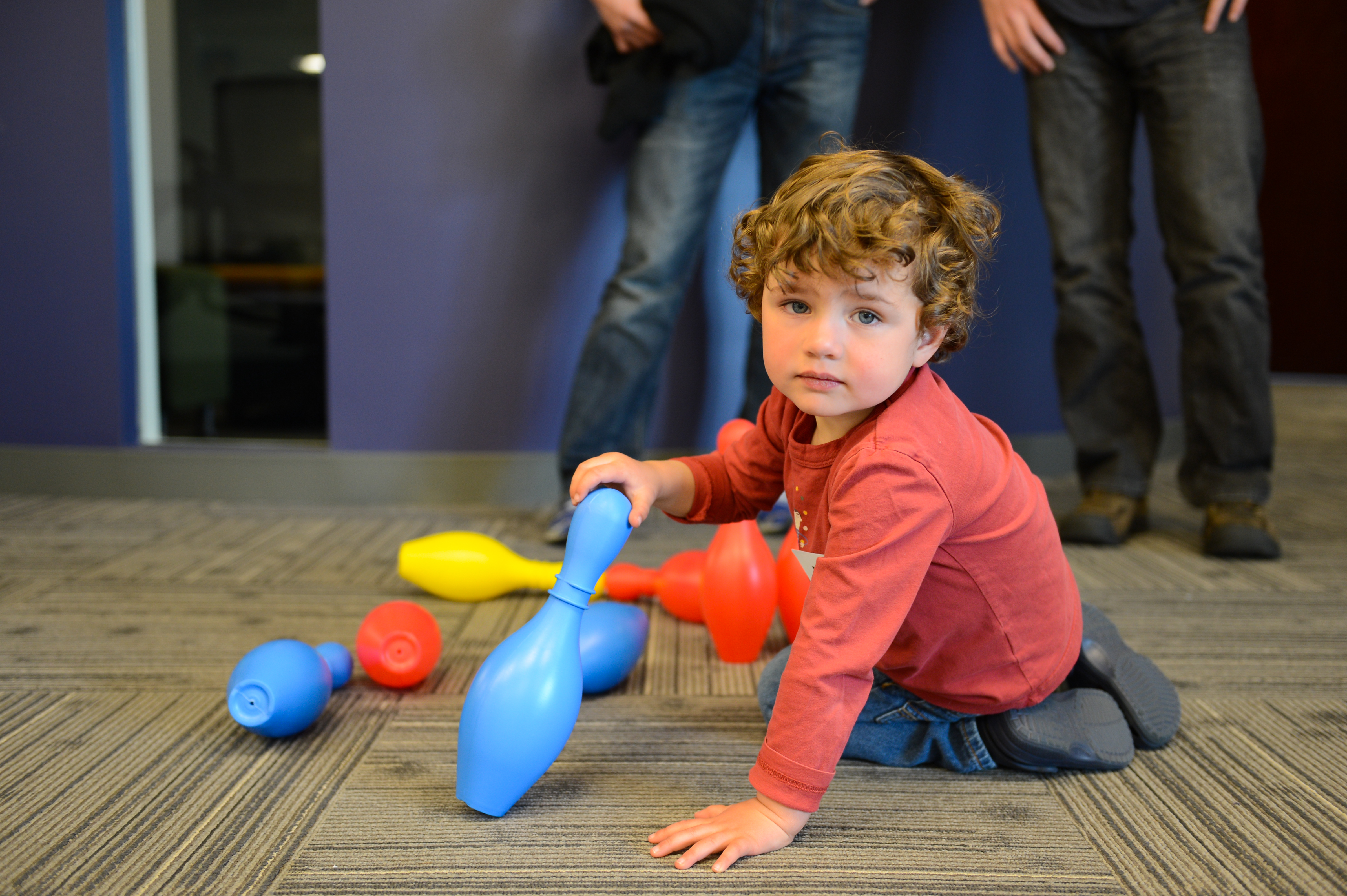 A young child in a red shirt plays with colorful plastic bowling pins on the floor.