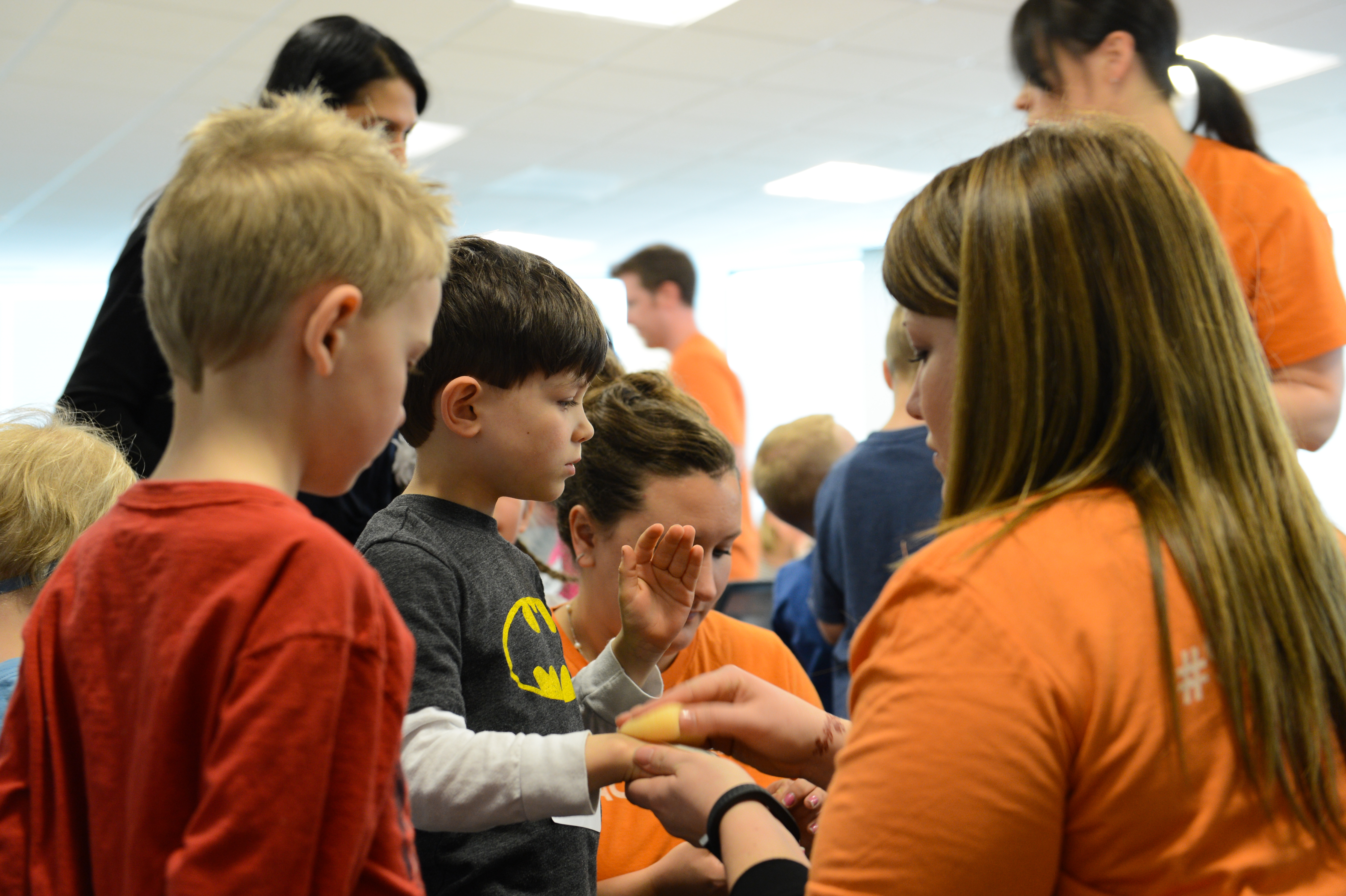 A woman in an orange shirt helps a child put on a wristband while another child watches.