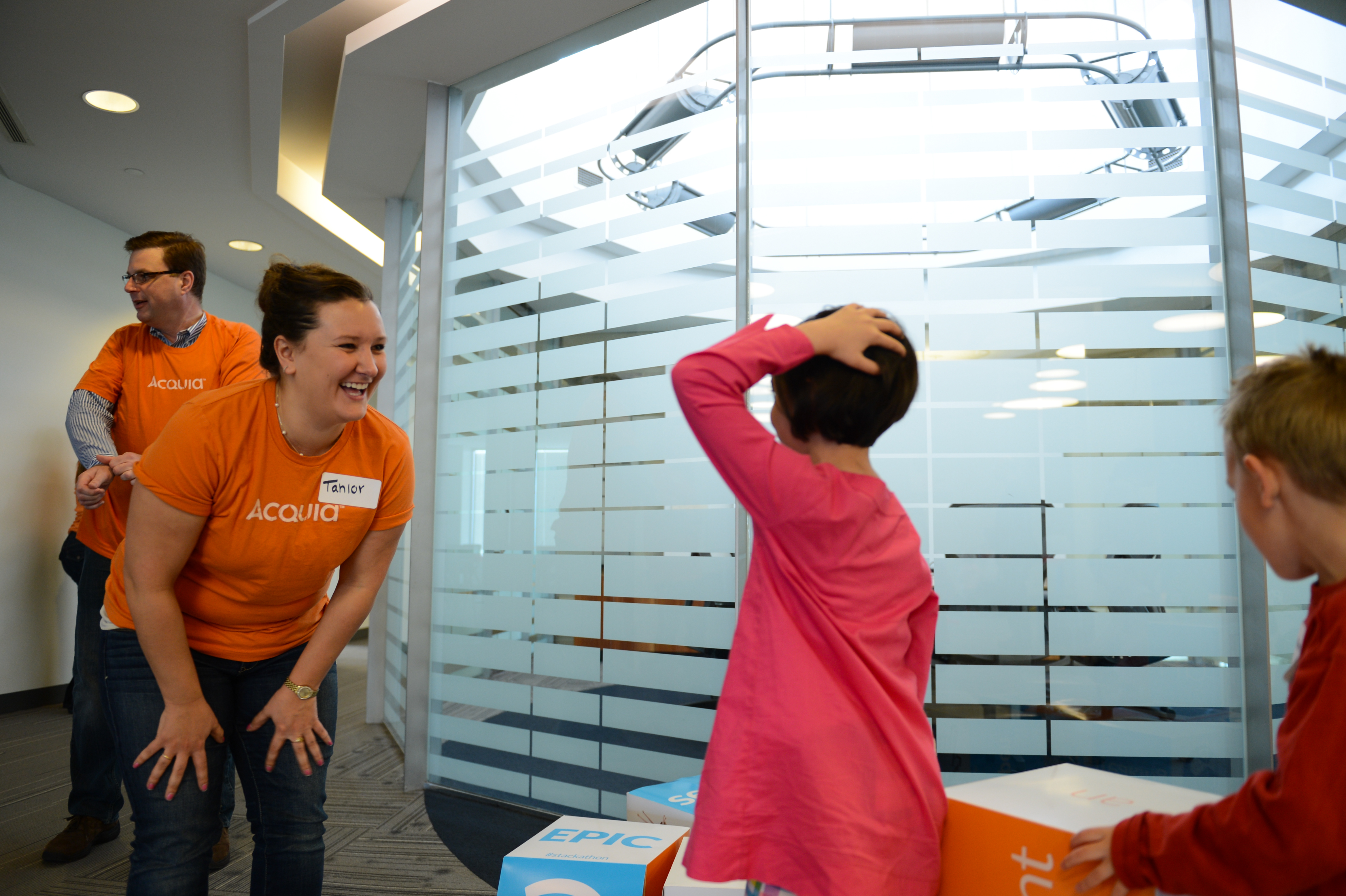 A woman in an orange Acquia shirt laughs while interacting with children playing with large blocks in an office.