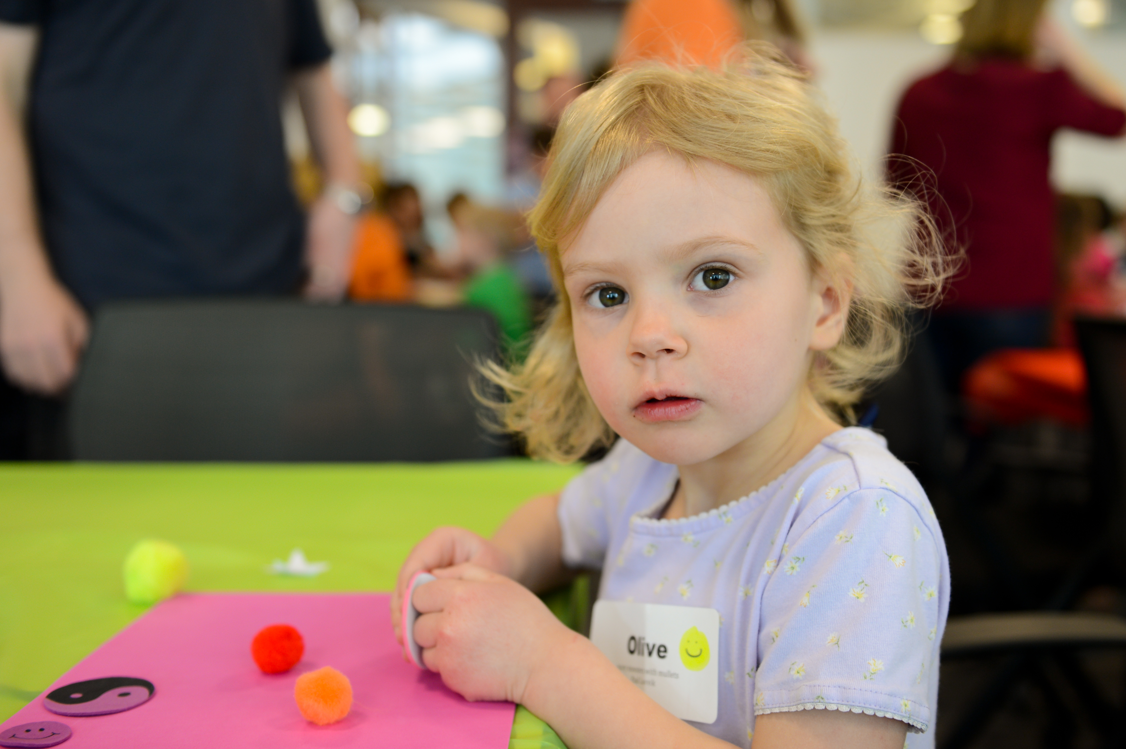 A young child sits at a table, working on a craft project with colorful pom-poms and paper.