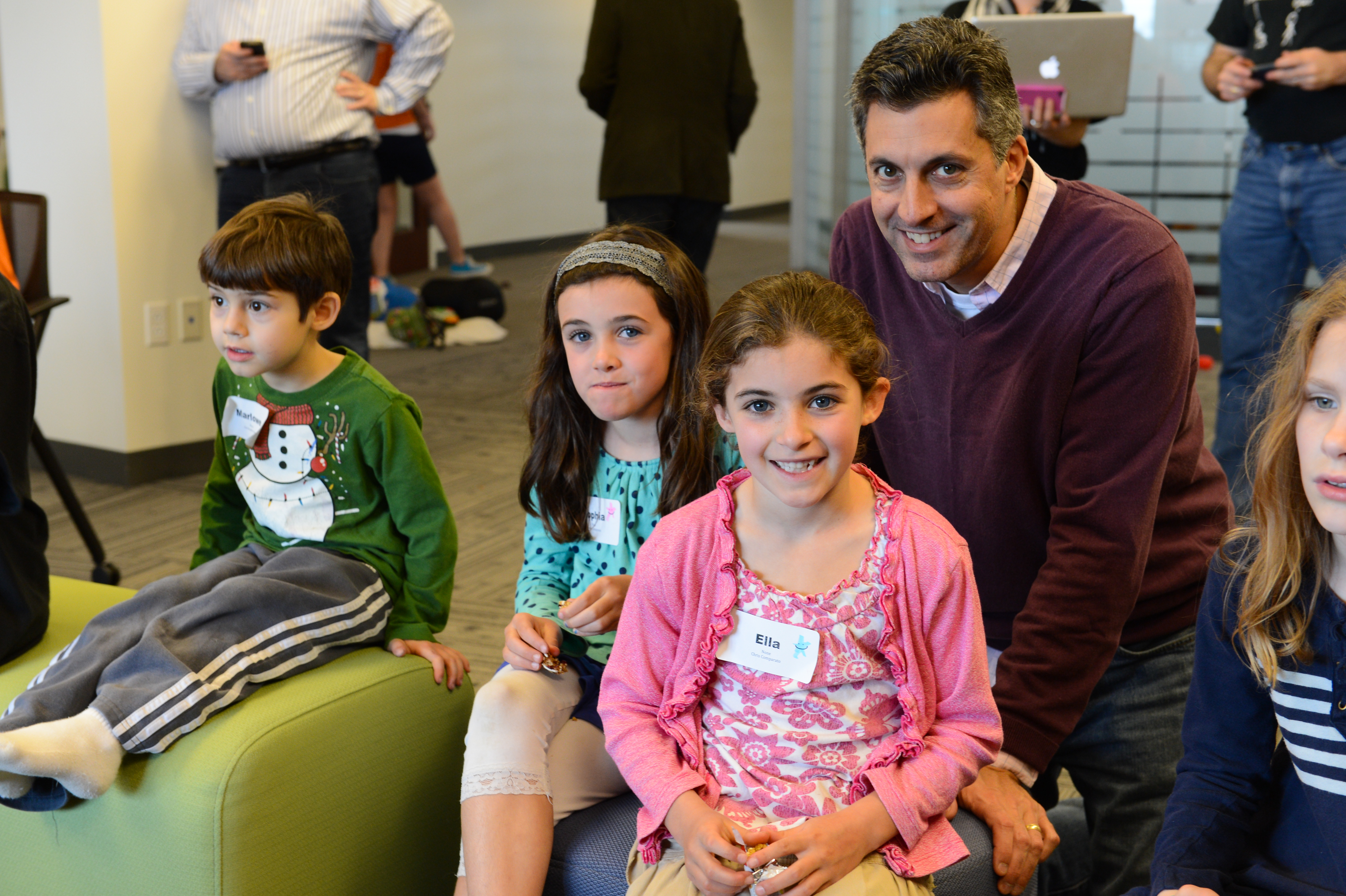 A man crouches next to smiling children in an office setting during a "Bring Your Kids to Work" event.