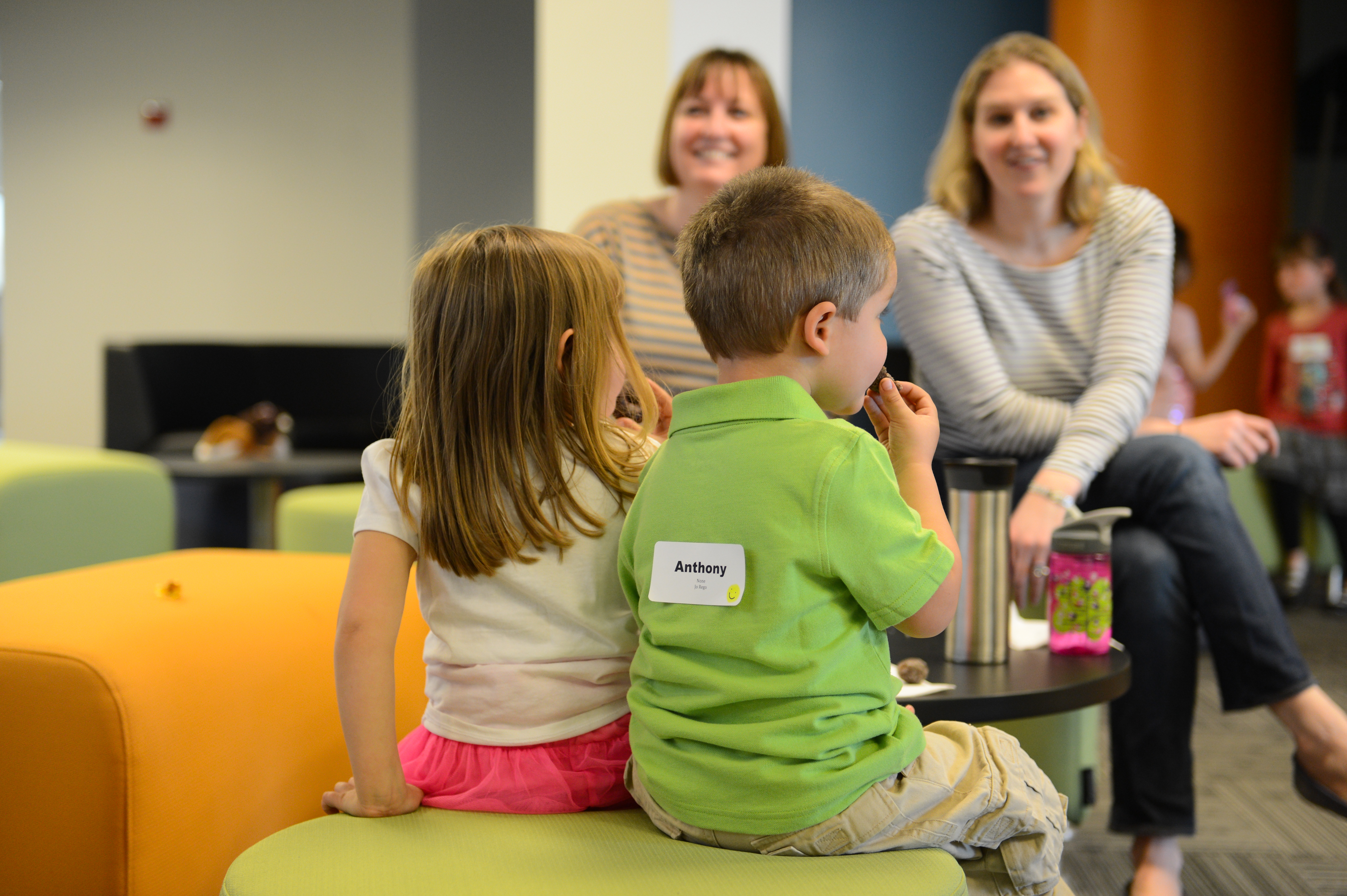 Two young children sit on a stool eating snacks, while two smiling adults watch in the background.