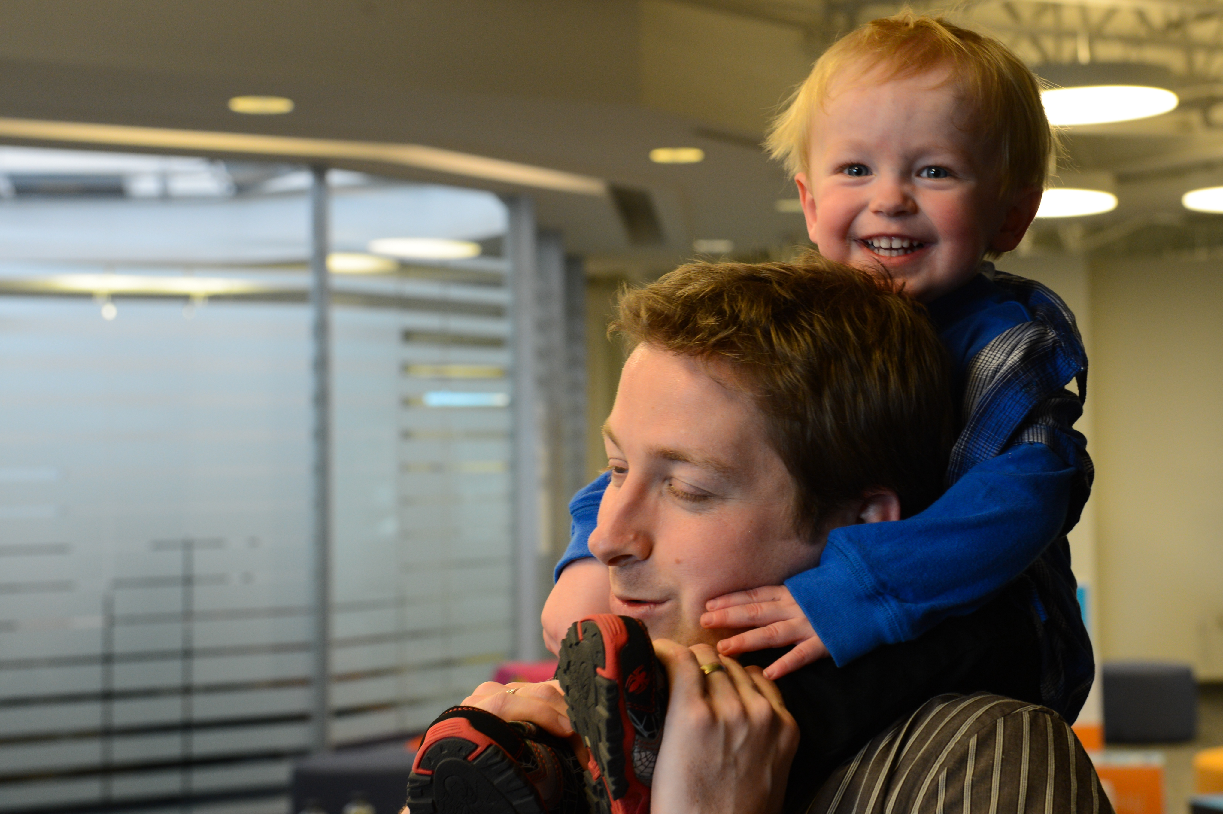 A smiling child in a blue sweater sits on an adult's shoulders, playfully holding onto their face.