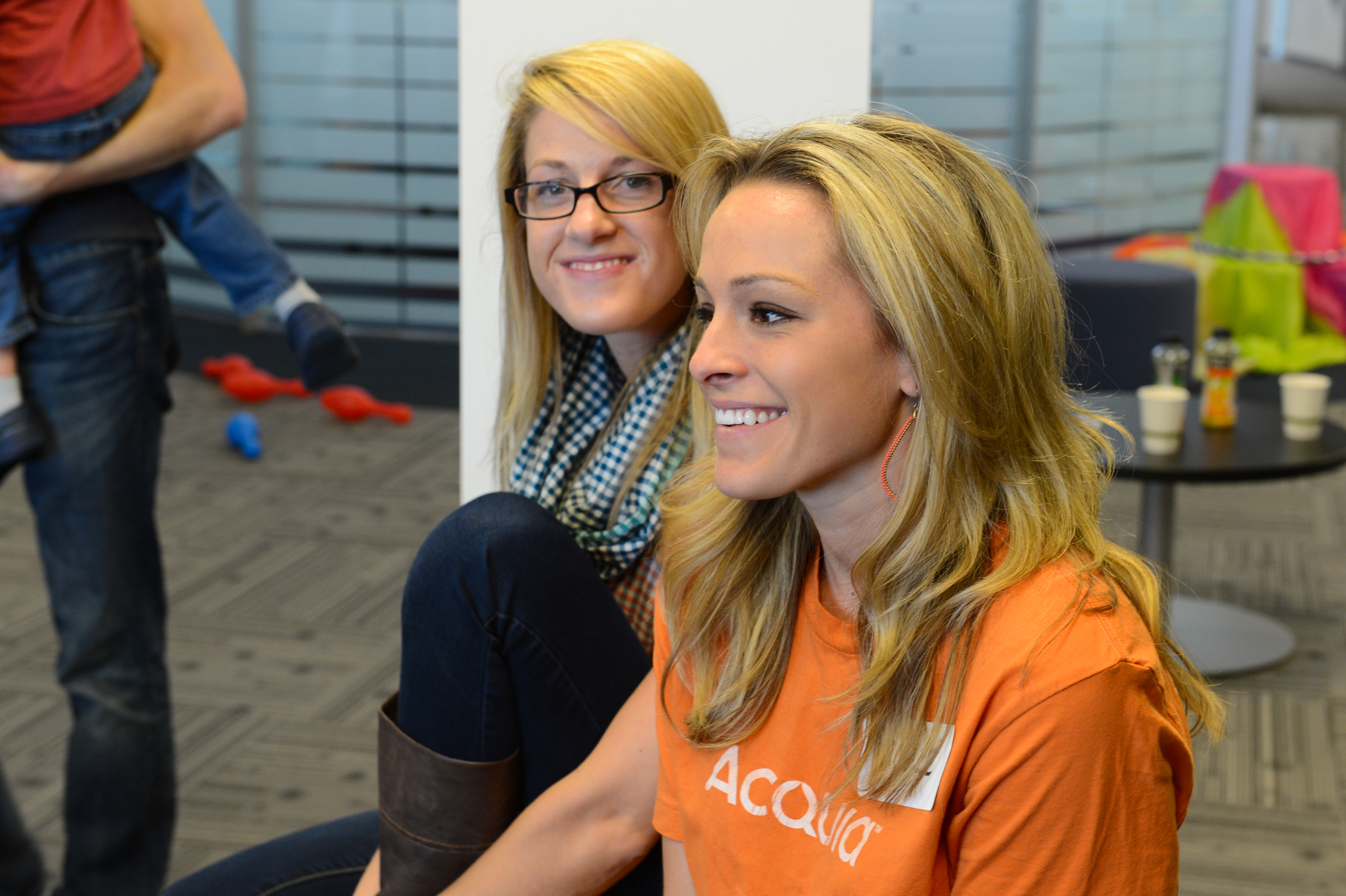 Two smiling women sit together in an office setting, one wearing an Acquia shirt during a company event.