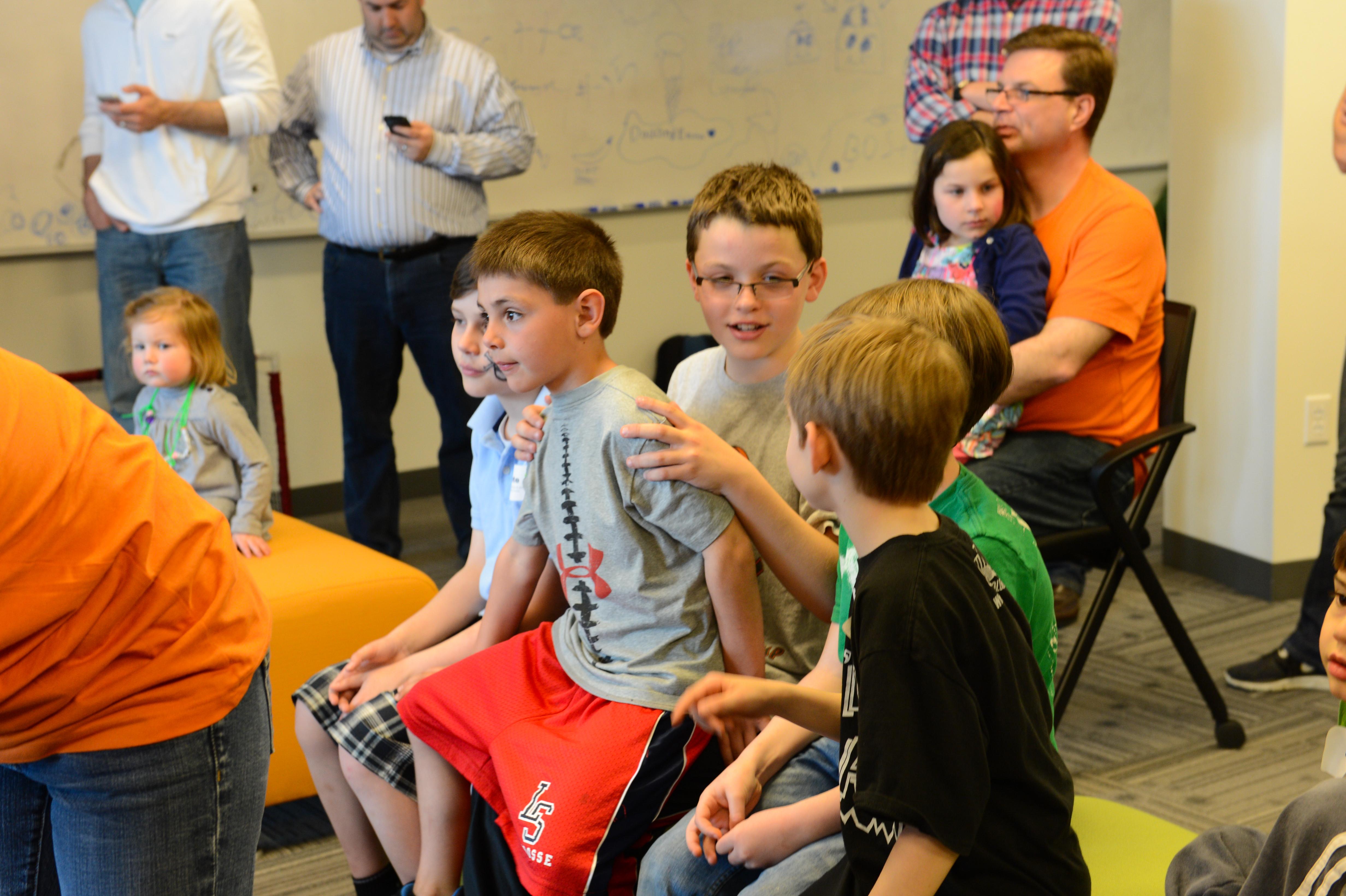 A group of children sits together, talking and interacting, while adults and other children observe in the background.