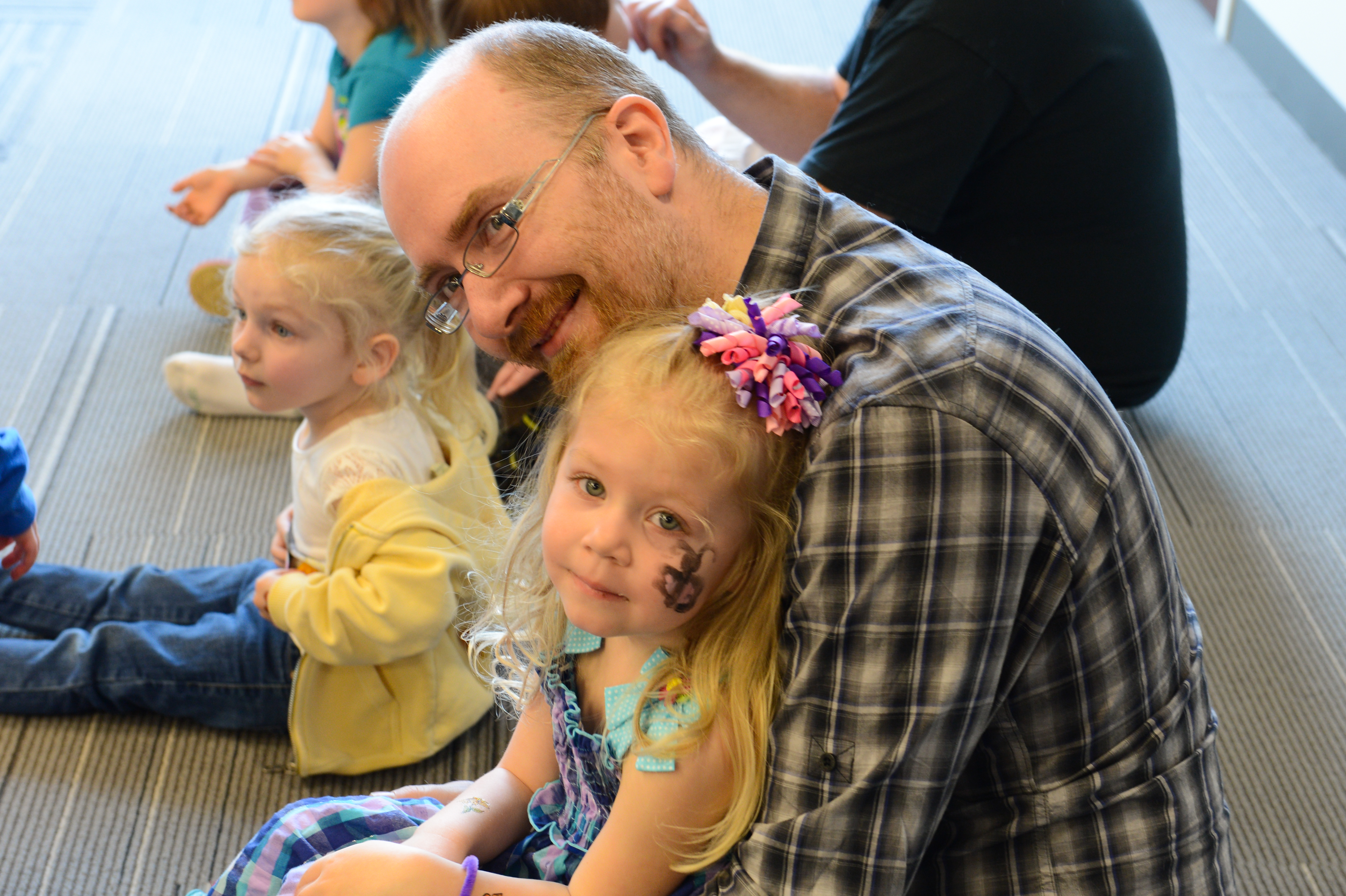 A man in glasses smiles while sitting with a young girl who has face paint and a colorful hair bow.
