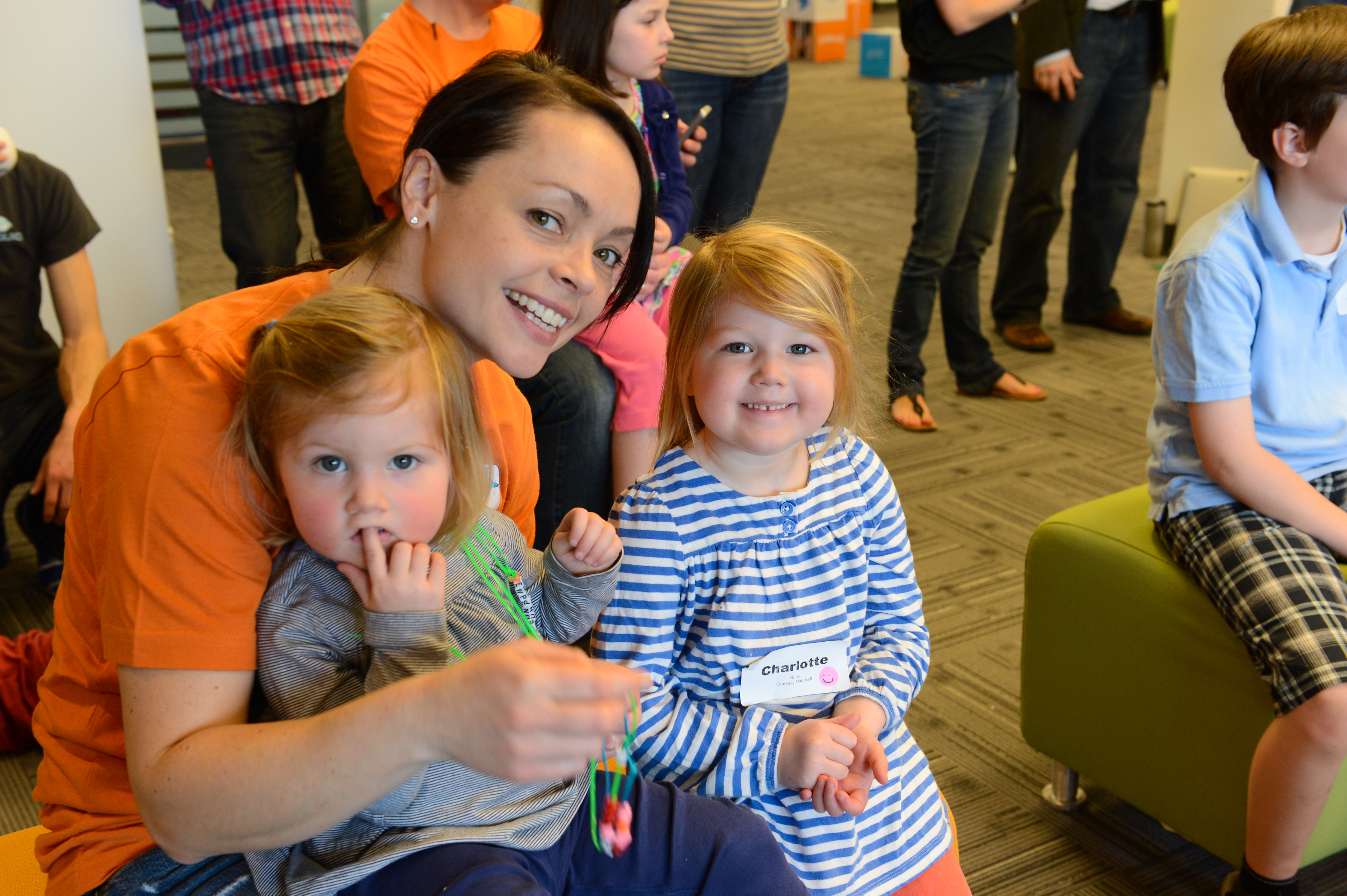 A smiling woman in an orange shirt sits with two young children, one holding beads, at a workplace event.