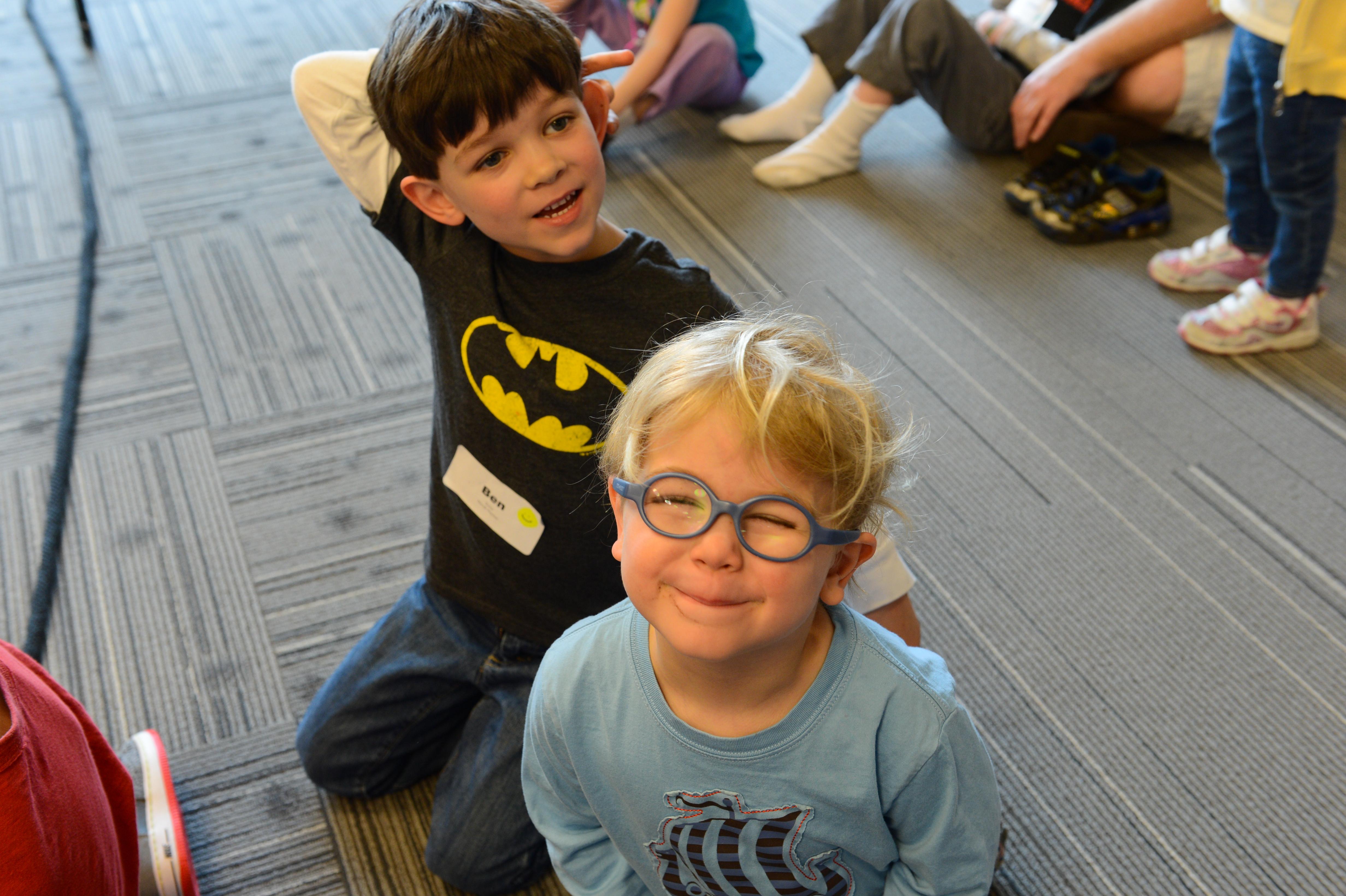 Two young children smile at the camera while sitting on the floor during a workplace event.