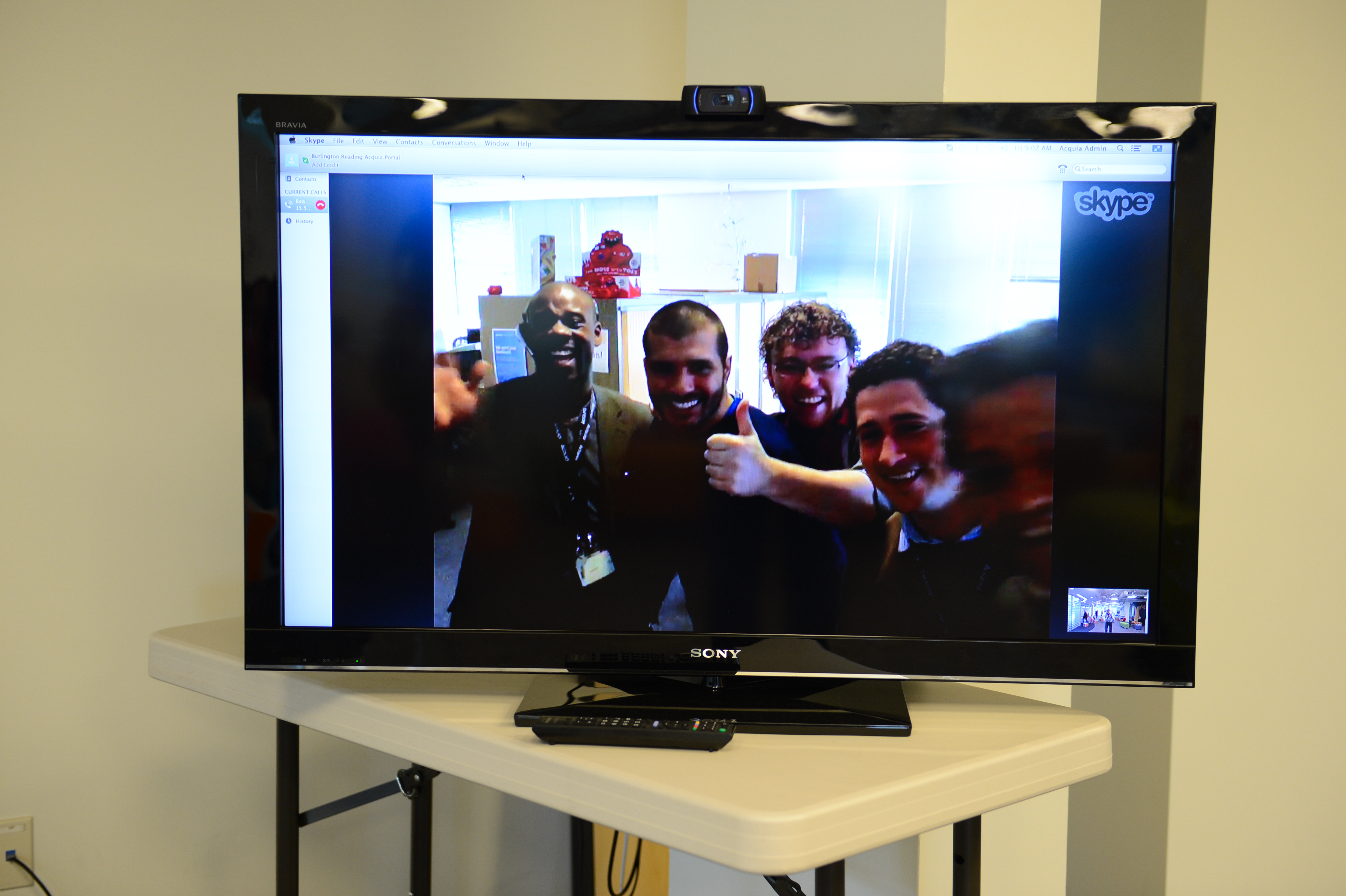 A television screen displays a video call with five smiling people waving and giving a thumbs-up.