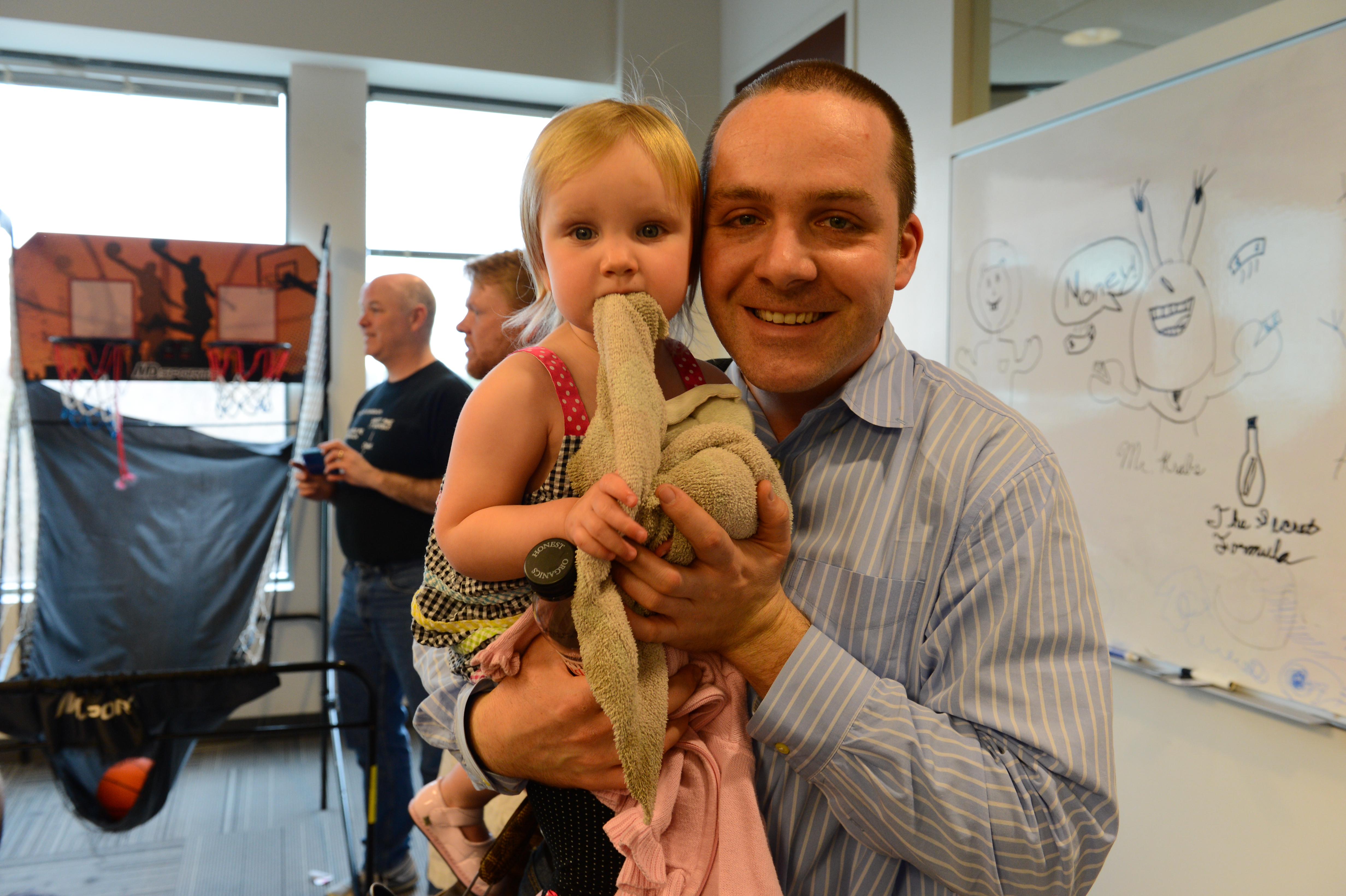 A smiling man holds a young child with a stuffed toy in an office setting.
