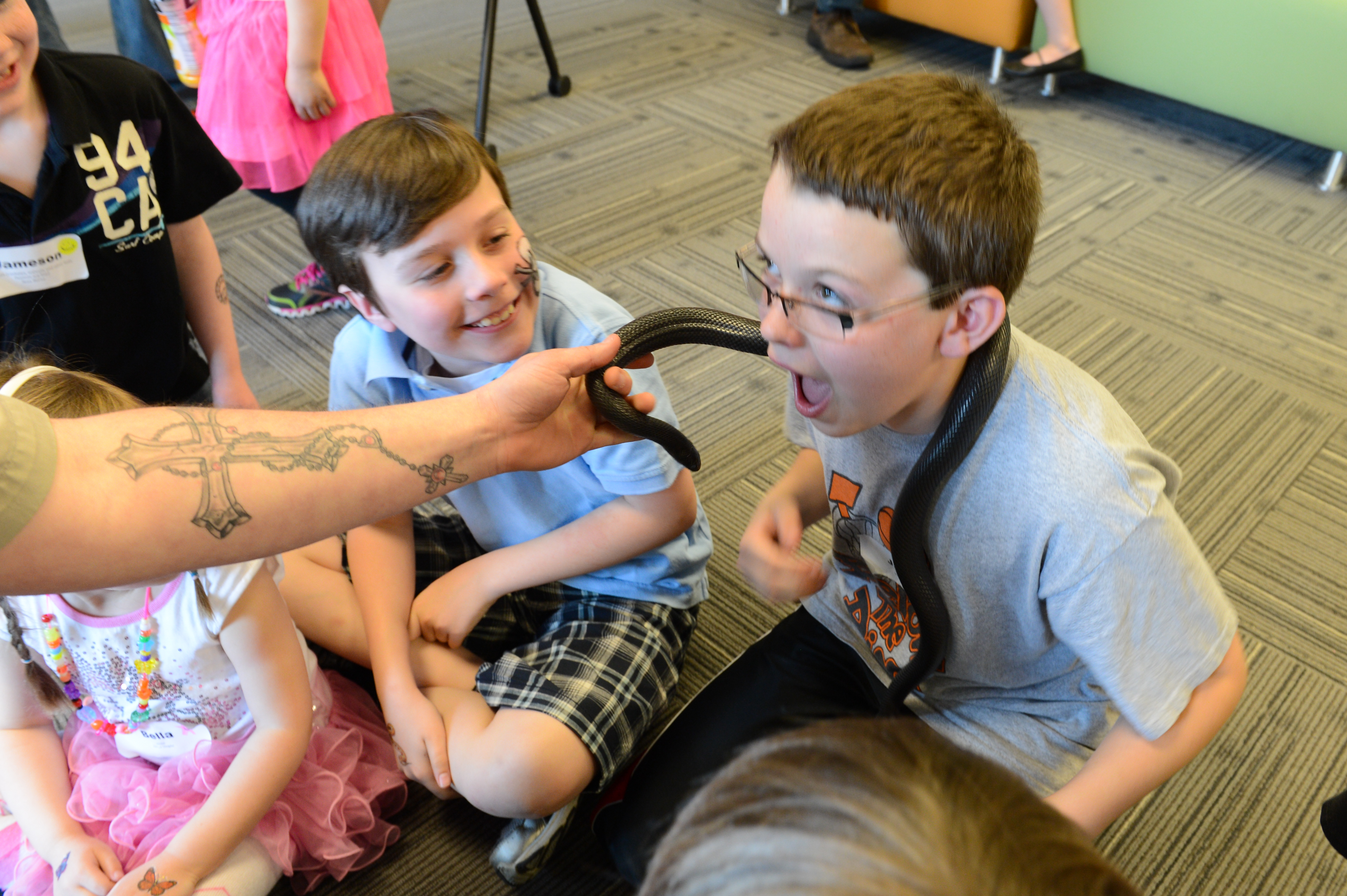 A child with glasses reacts excitedly as a snake is held near their face, while others watch and smile.