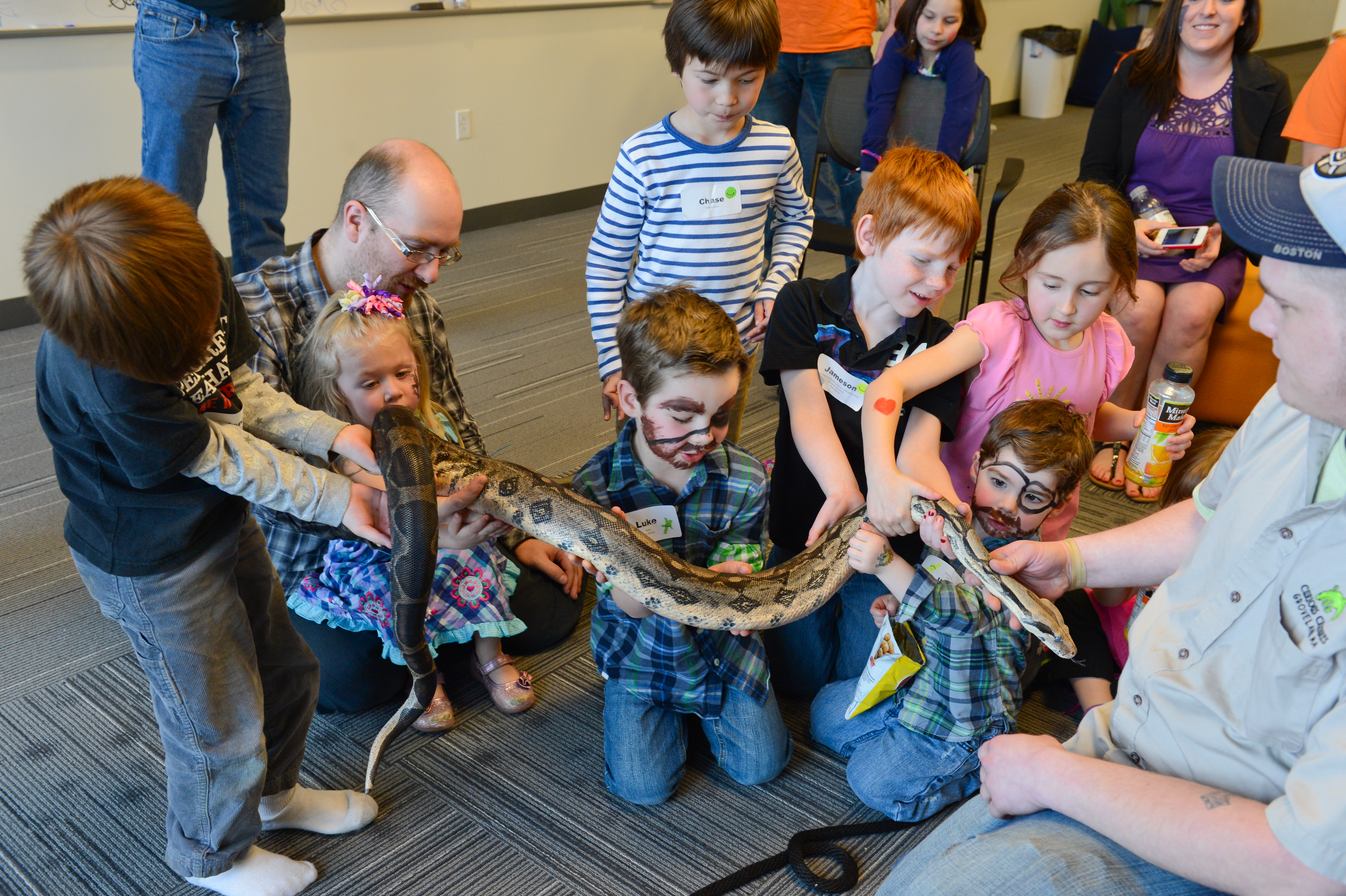 A group of children excitedly touch a large snake held by an animal handler during a workplace event.