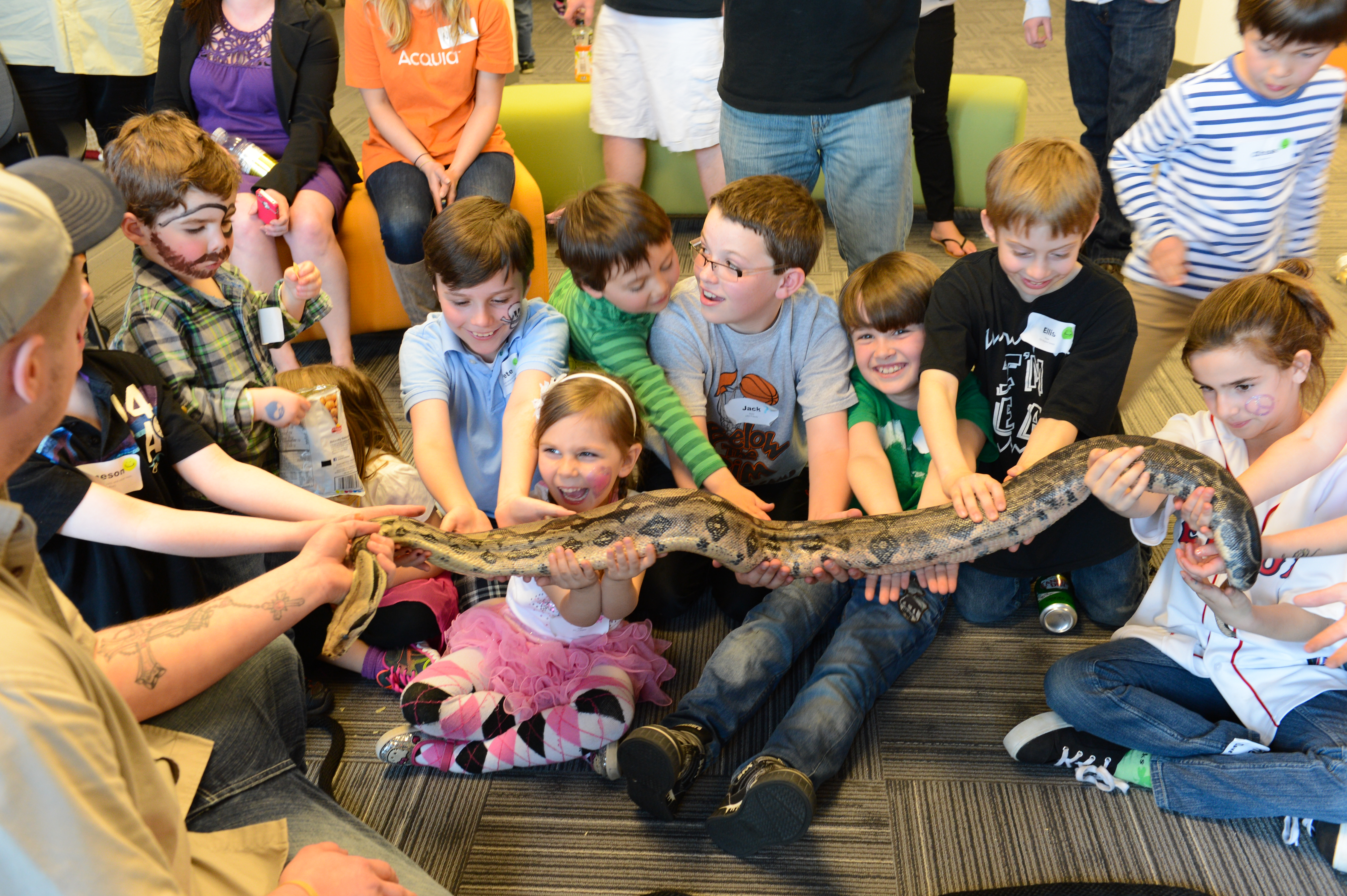 A group of children excitedly hold a large snake with the help of an adult during an event.