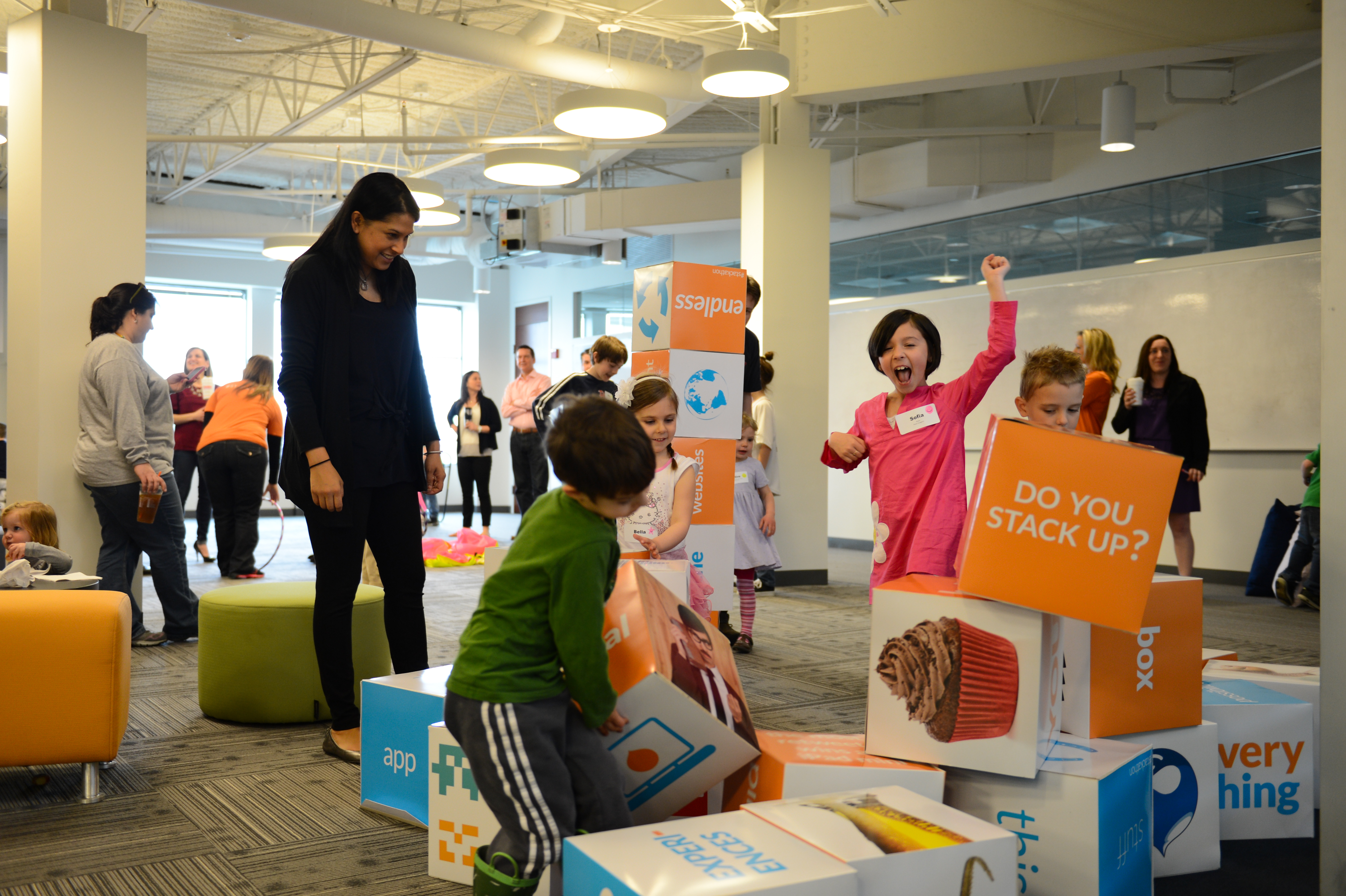 Children excitedly stack and knock over large colorful blocks while an adult watches in an office setting.