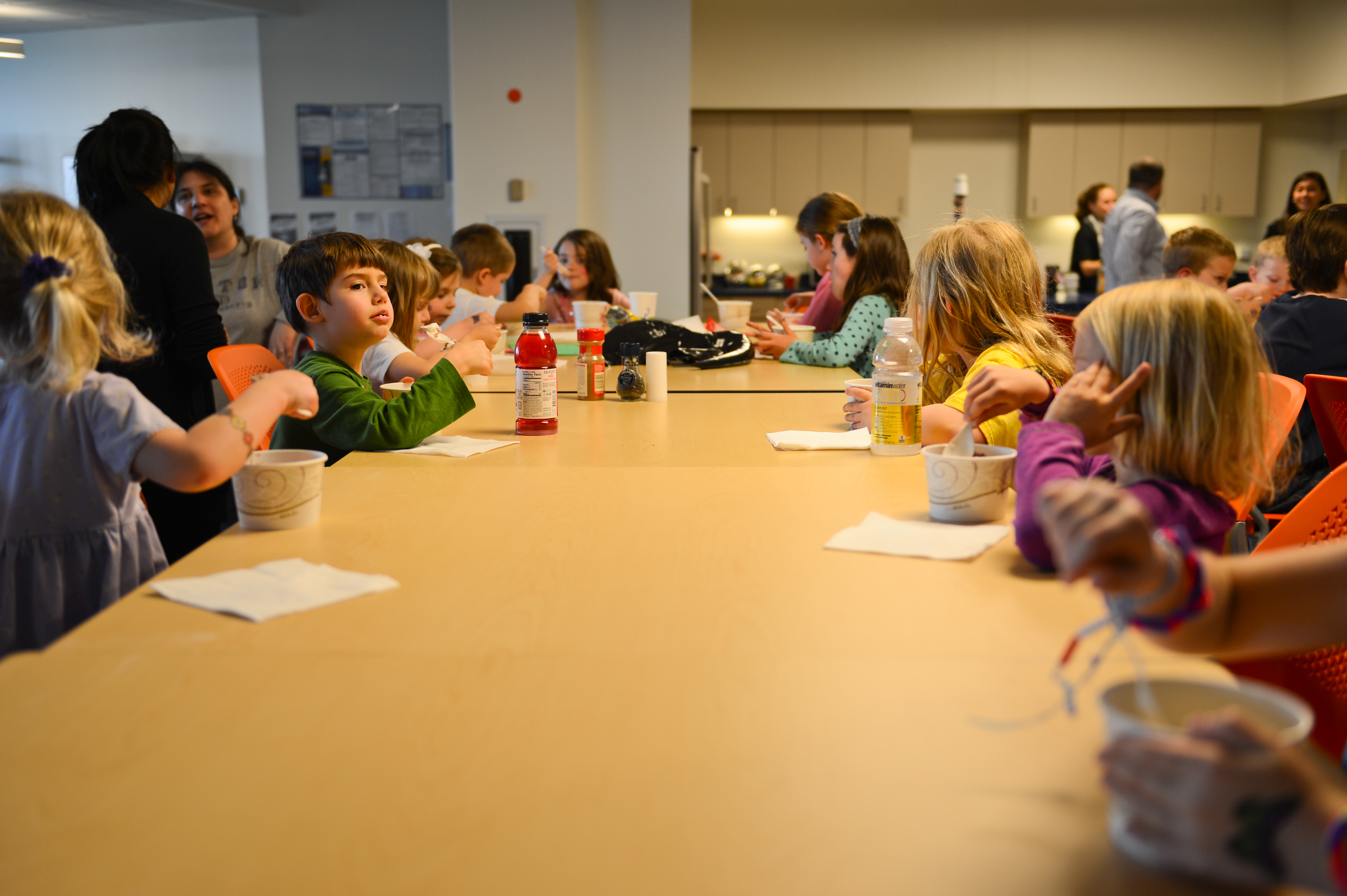Children sit around a long table, eating and talking during a "Bring Your Kids to Work Day" event.