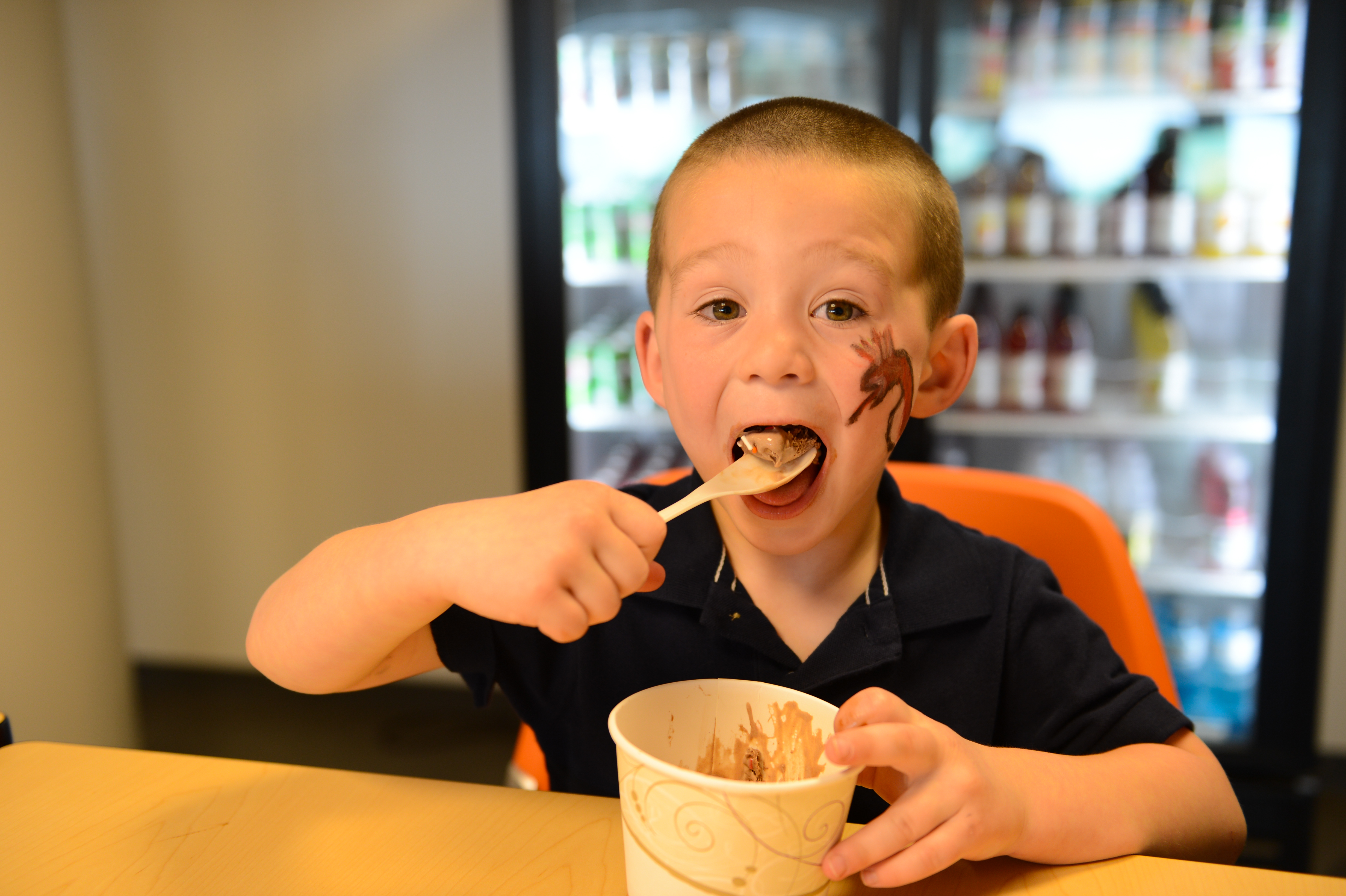 A young child with face paint eats chocolate ice cream with a wooden spoon at a table.