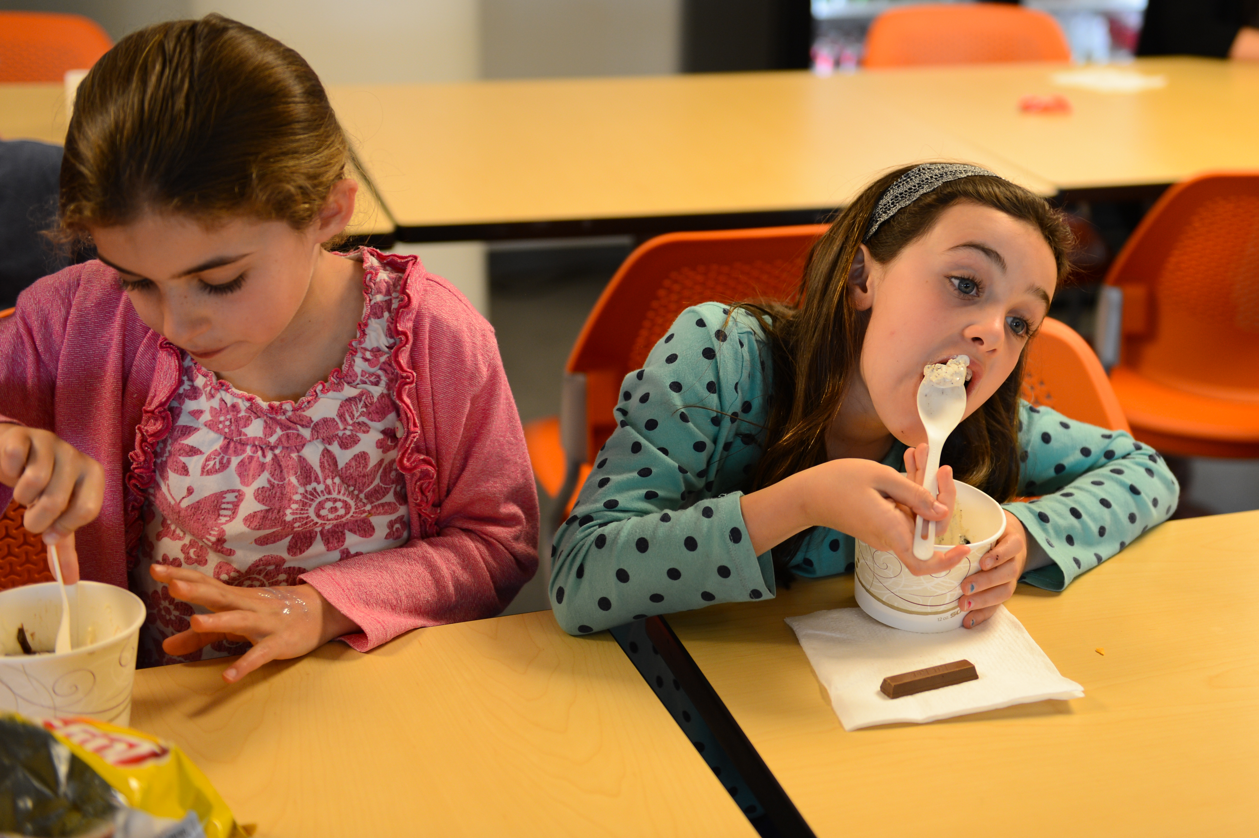Two children sit at a table eating from cups with spoons, one leaning forward while taking a bite.