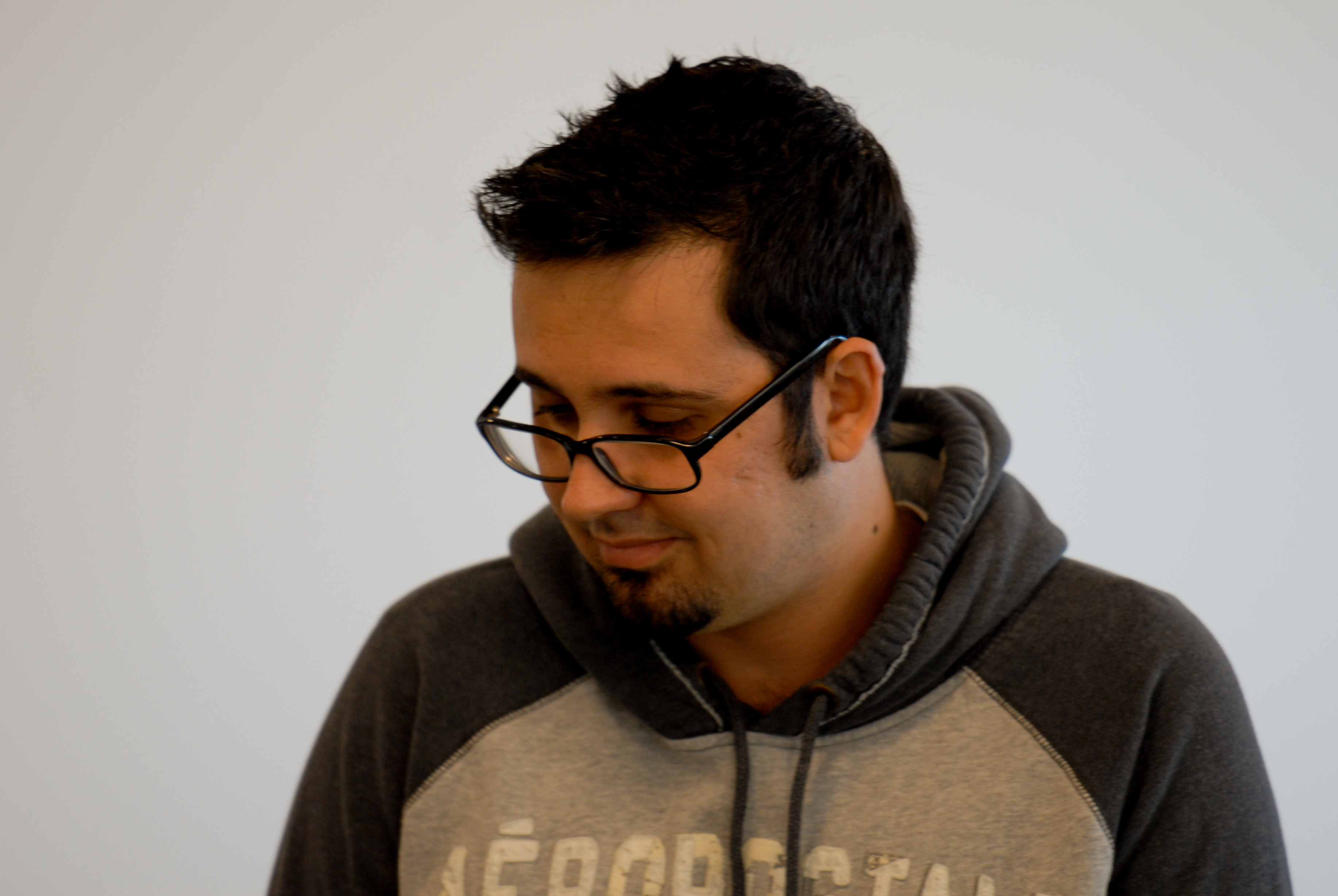 A person wearing glasses and a hoodie looks down with a slight smile in an indoor setting.