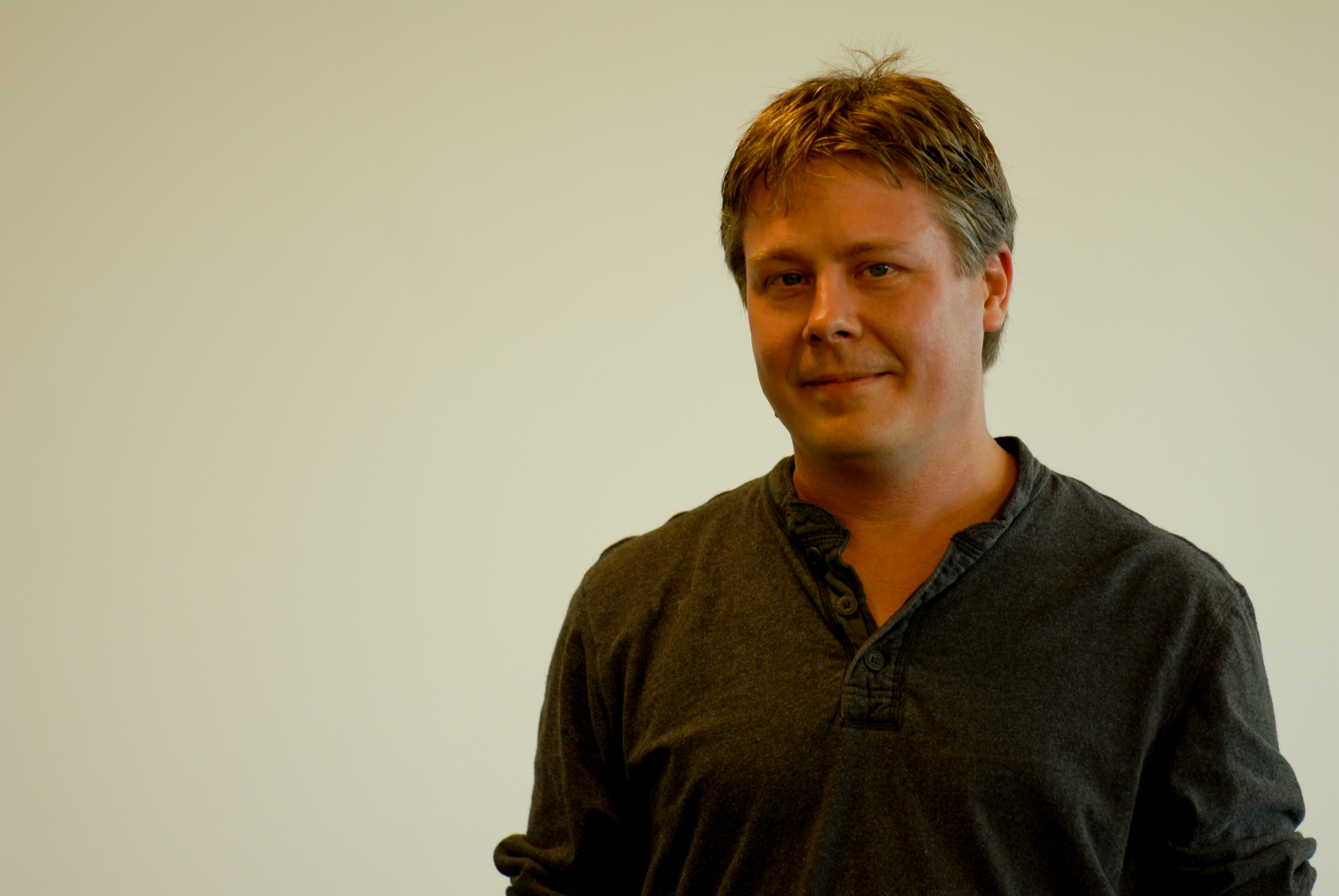A person in a dark shirt stands in front of a plain background, looking at the camera with a slight smile.