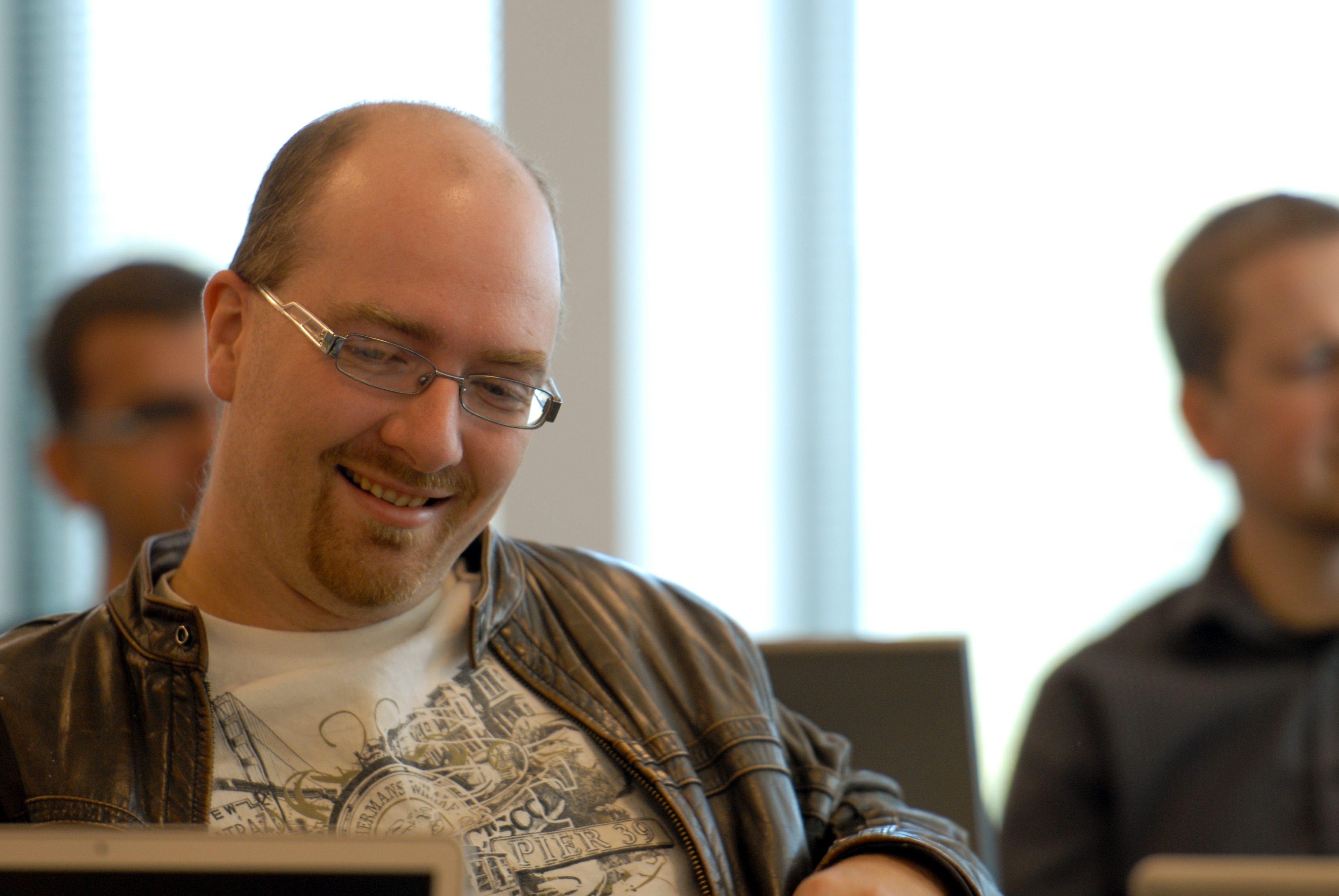 A man wearing glasses and a leather jacket smiles while looking at a laptop during a meeting or event.