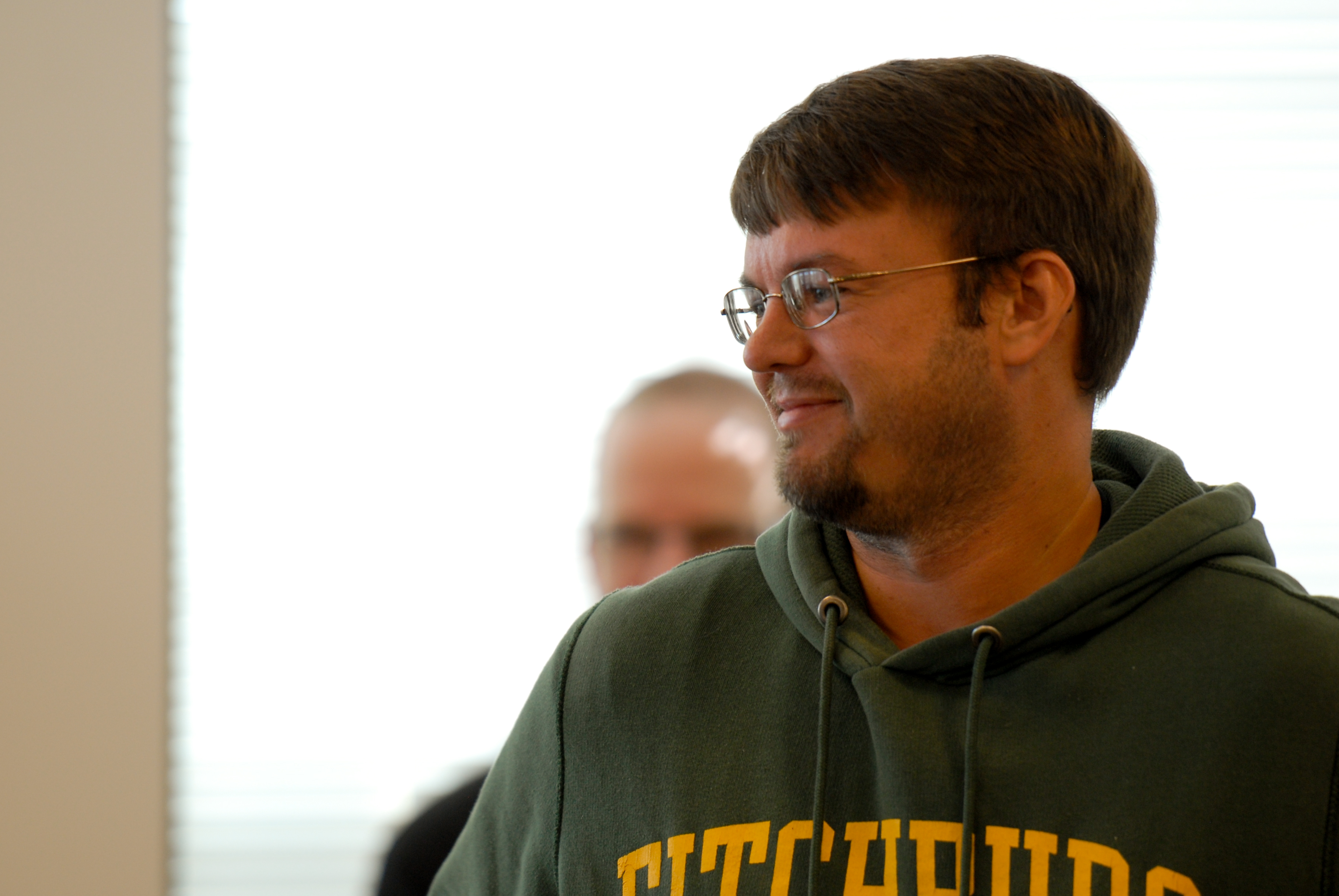 A man in a green hoodie and glasses smiles while looking to the side, with another person blurred behind him.
