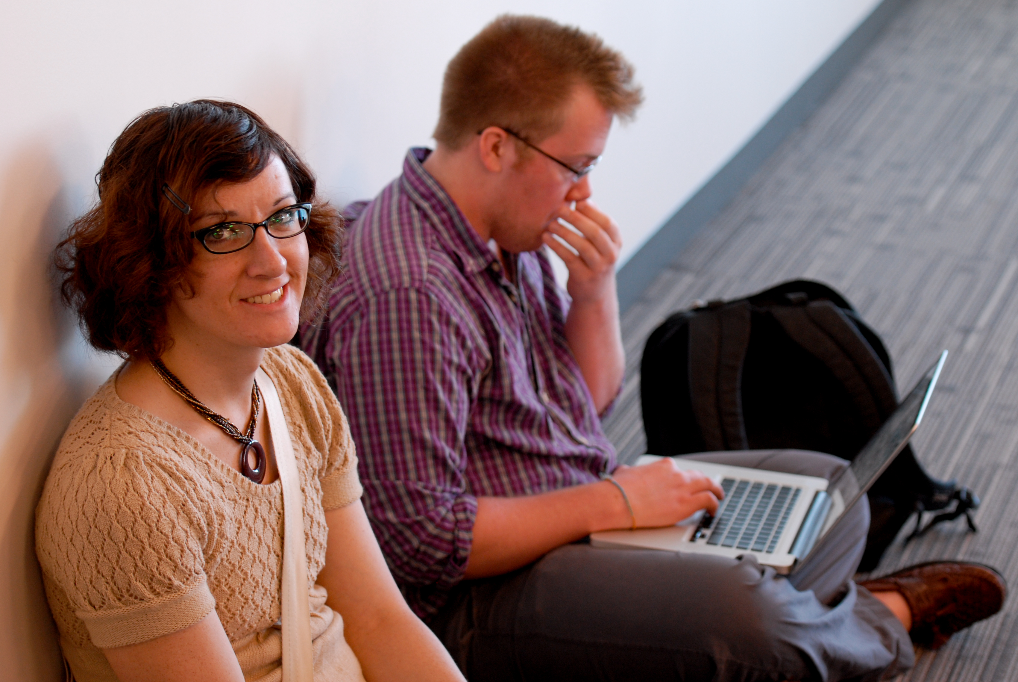 A woman smiles at the camera while a man sits beside her, focused on his laptop.