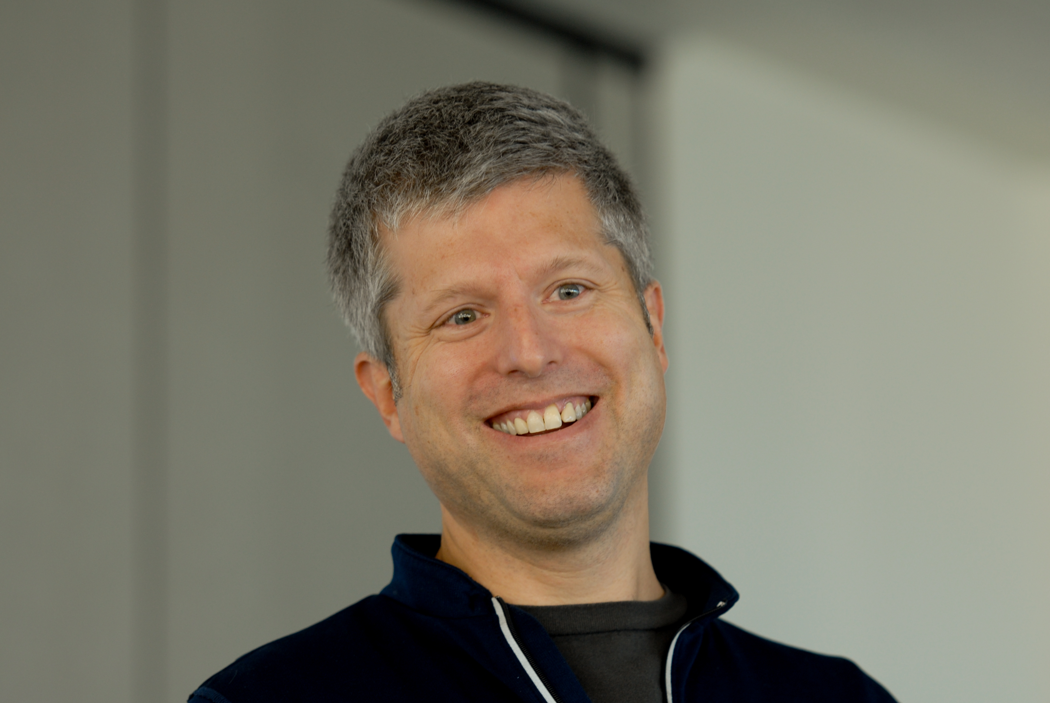 A smiling person with short gray hair wearing a dark jacket, looking slightly off-camera in an indoor setting.
