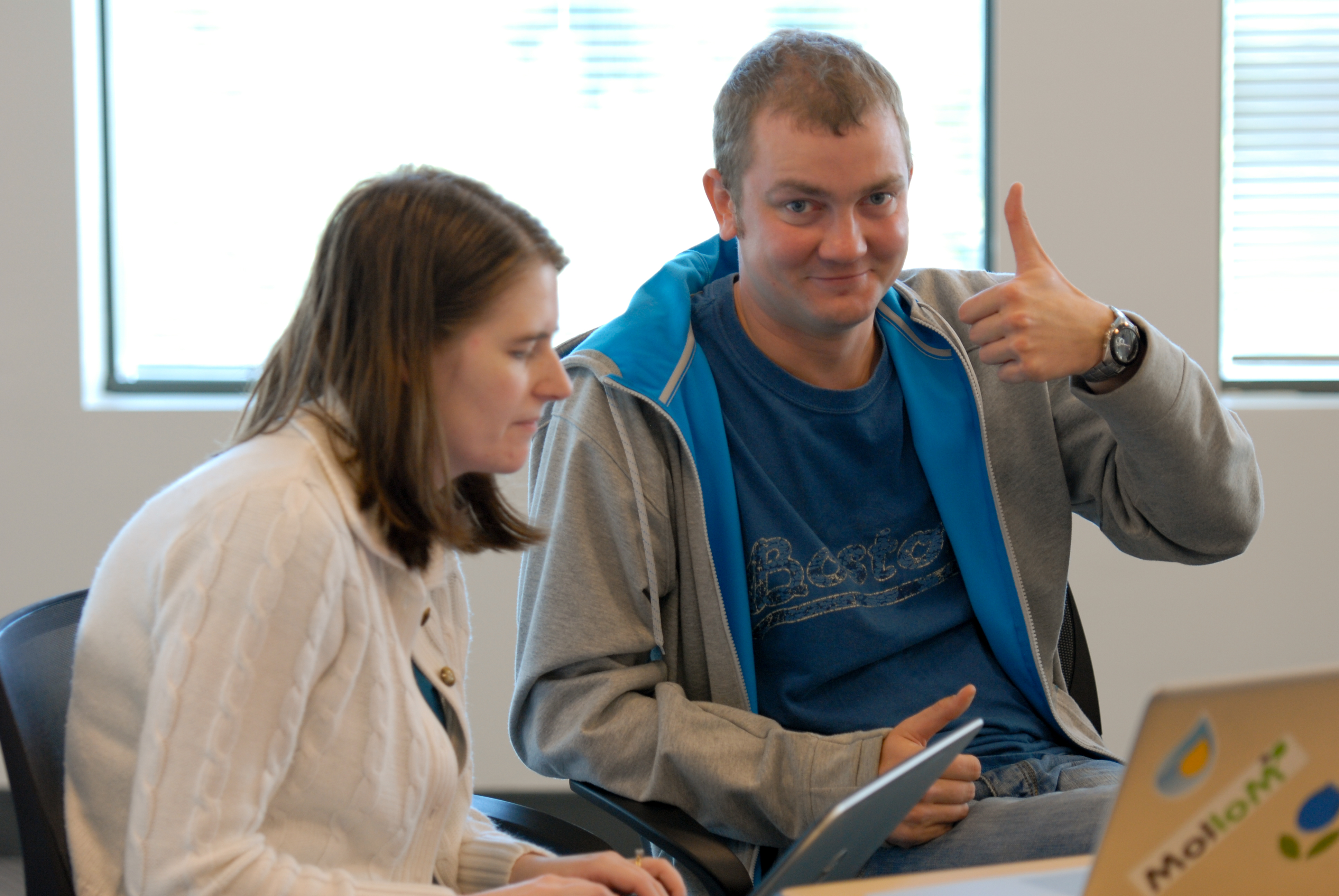 A woman focuses on her laptop while a man beside her gives a thumbs-up and smiles.