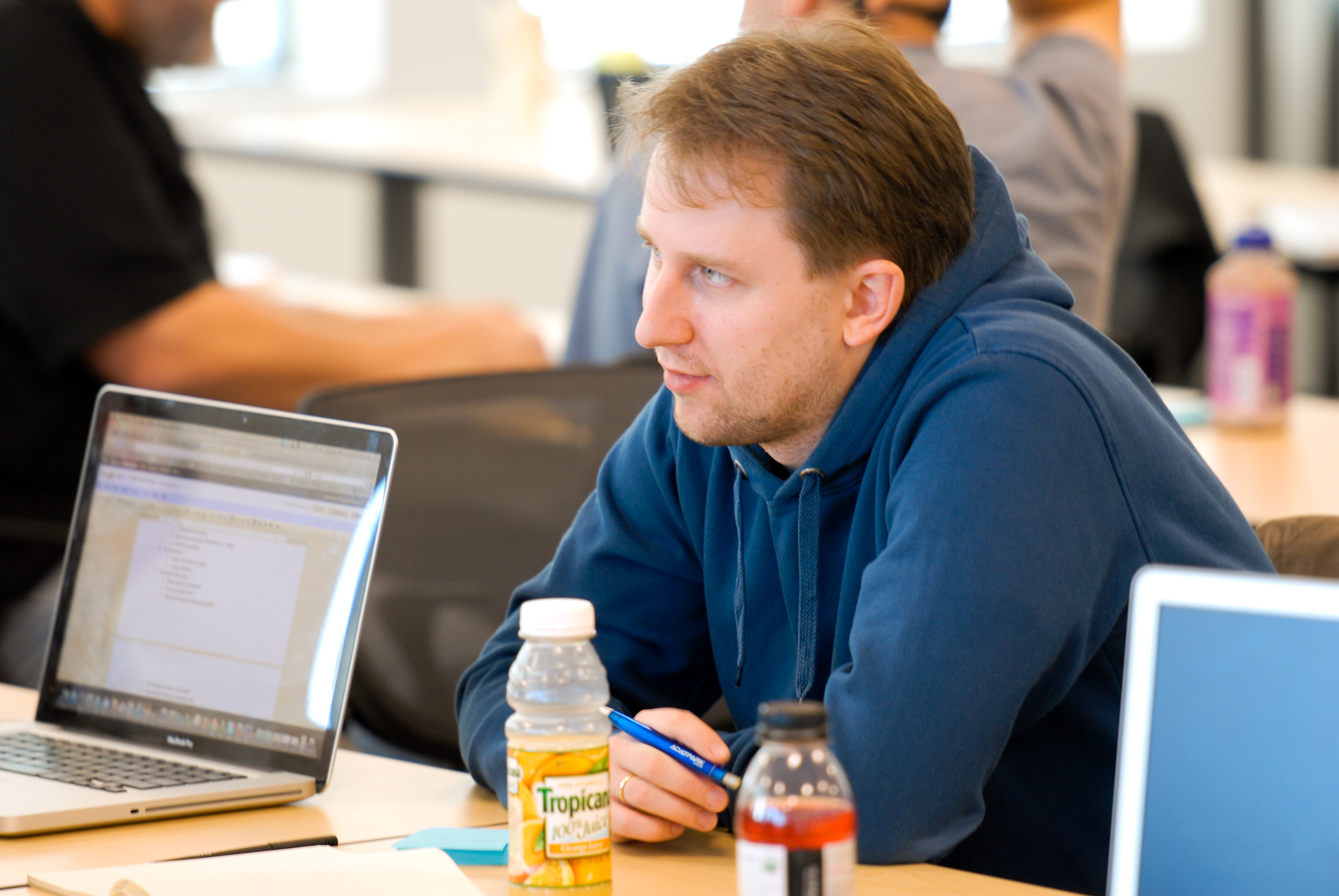 A man in a blue hoodie listens attentively during a meeting, holding a pen near an open laptop.