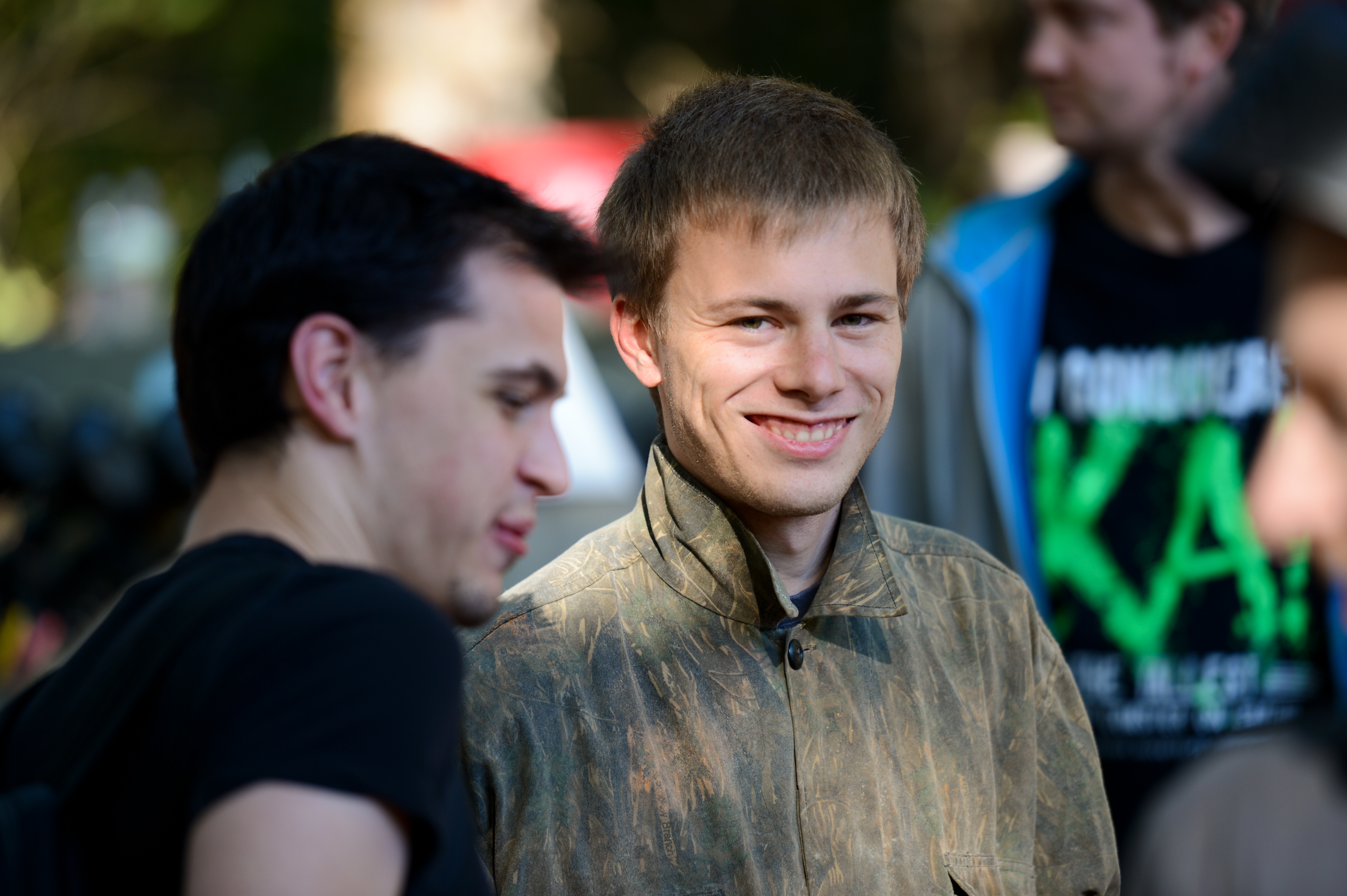A smiling person in a brown jacket looks at the camera while another person speaks nearby in an outdoor setting.