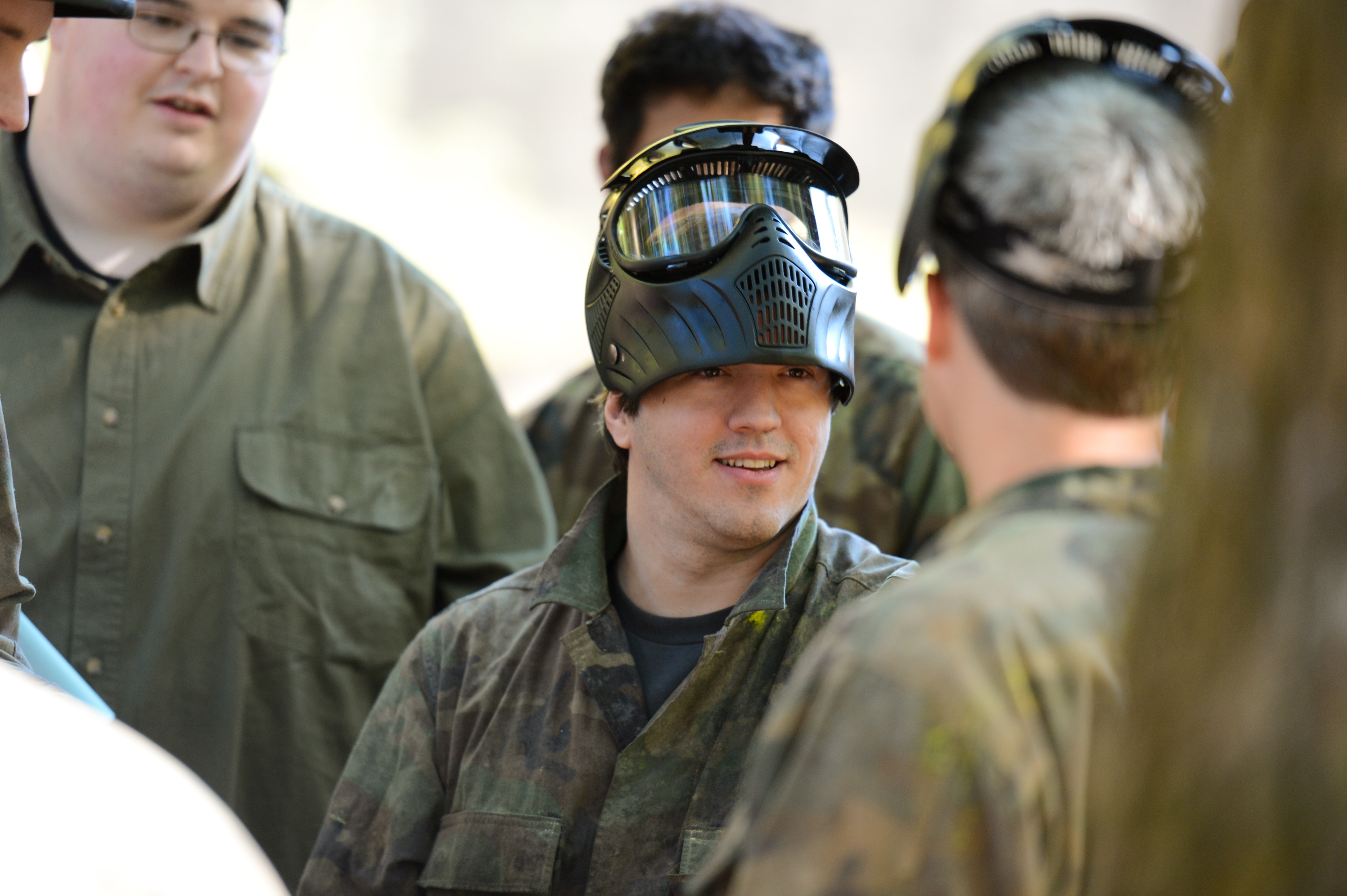 A group of people in camouflage clothing and paintball masks talk before a game.