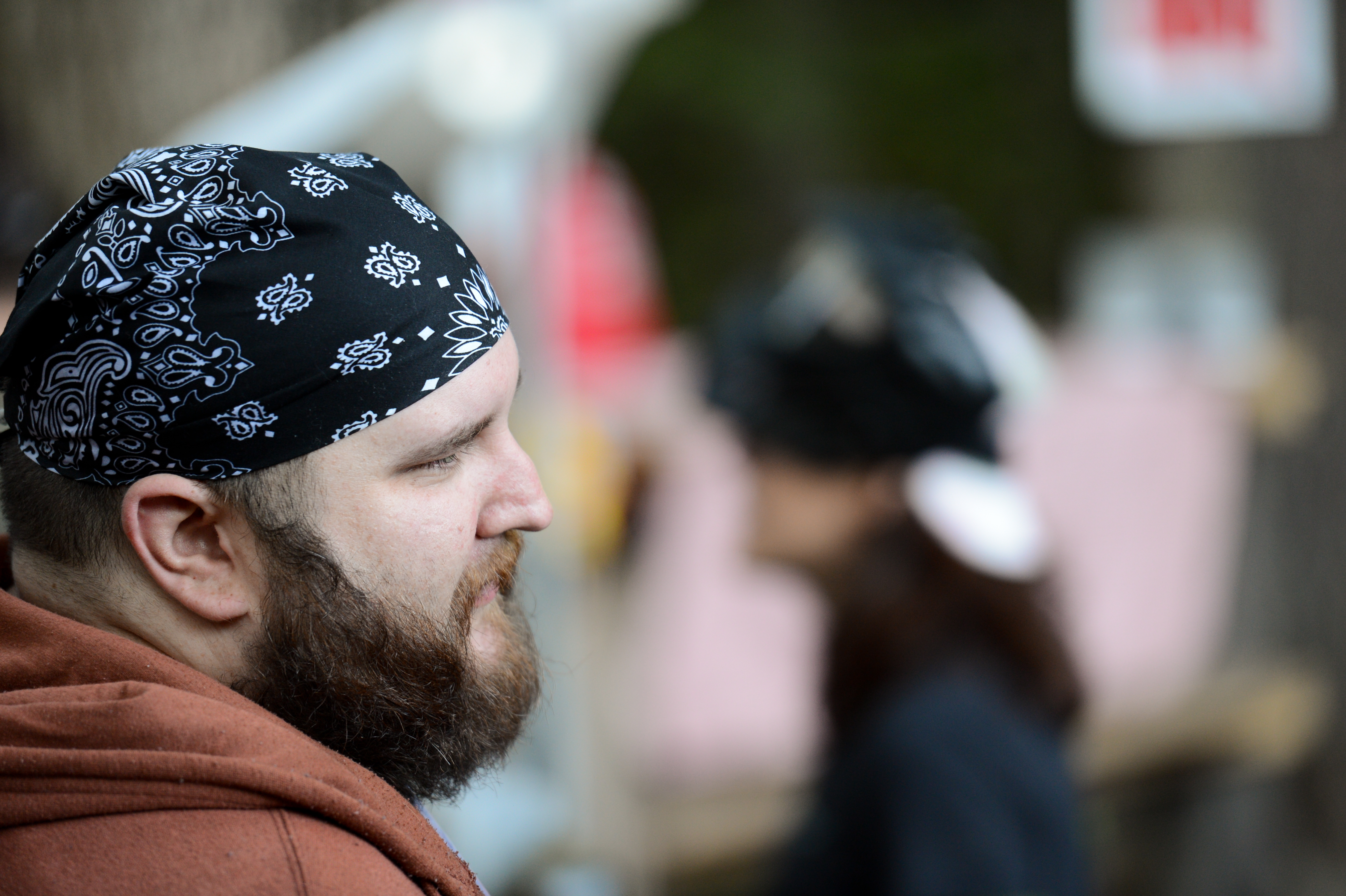 A bearded person wearing a black bandana looks to the side, with another person blurred in the background.